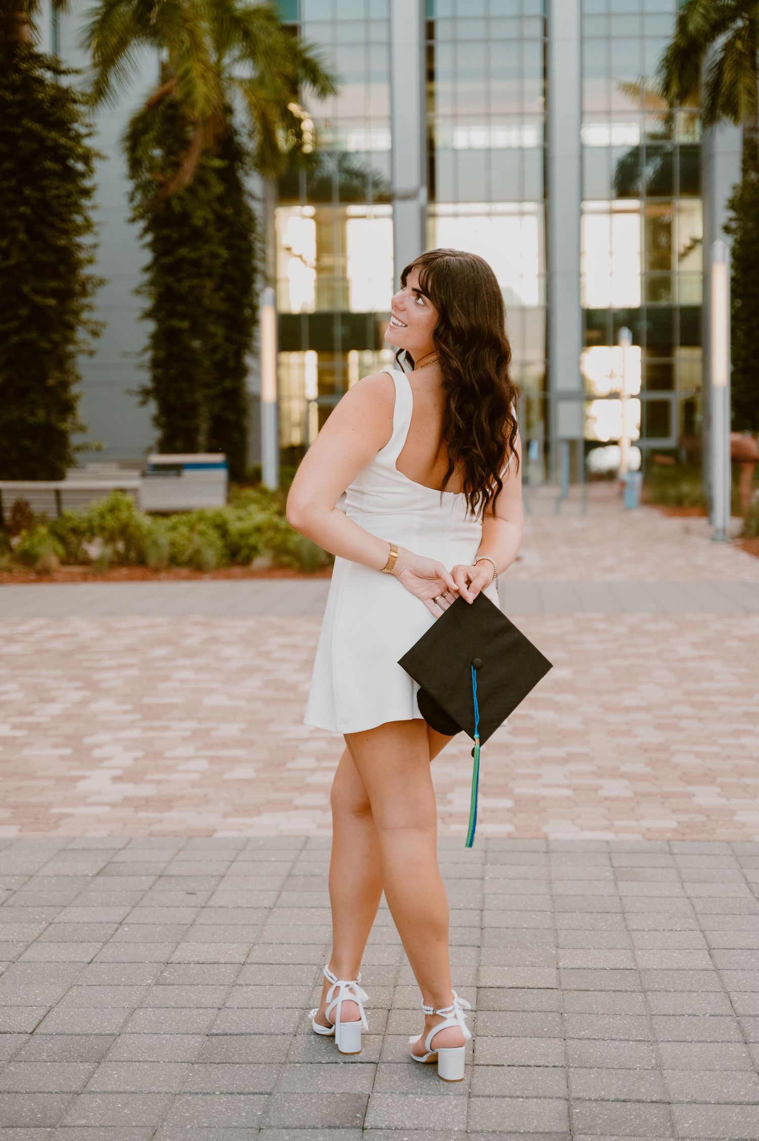 Graduate in white dress poses playfully with graduation cap outside modern building.