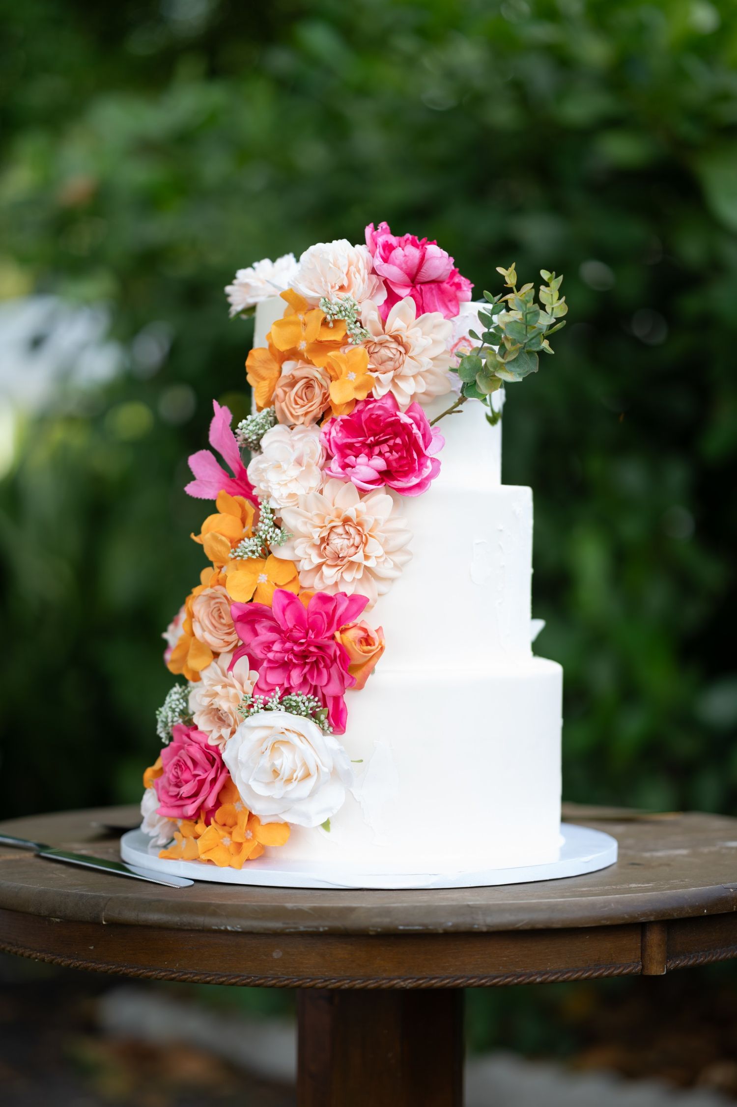 White wedding cake decorated with cascading pink and orange flowers on wooden stand.