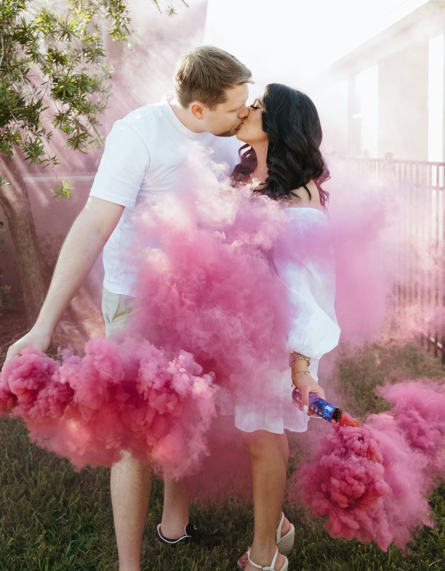 Romantic couple sharing a moment surrounded by pink smoke bombs in a backyard.