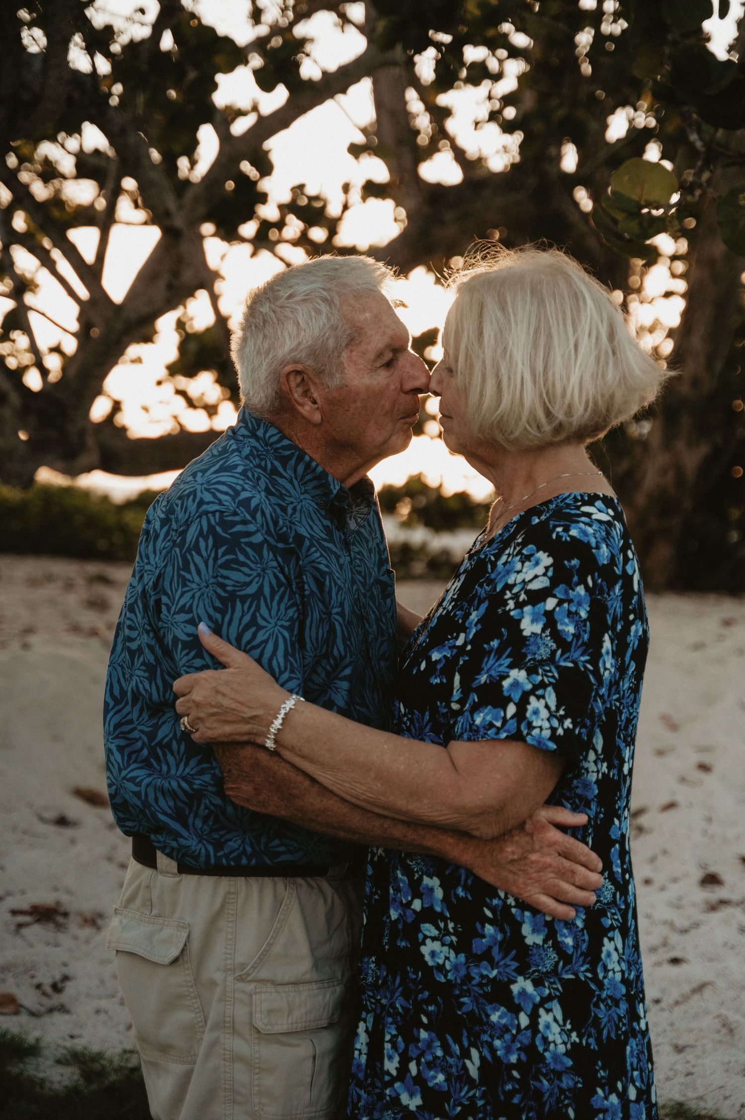 Elderly couple embracing on the beach at sunset wearing blue and khaki clothing.