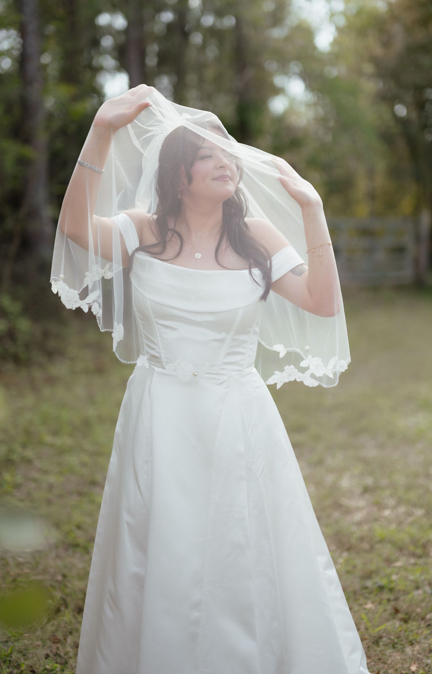 A bride in a white wedding dress and veil poses outdoors, lifting her veil with both hands in a serene garden setting.