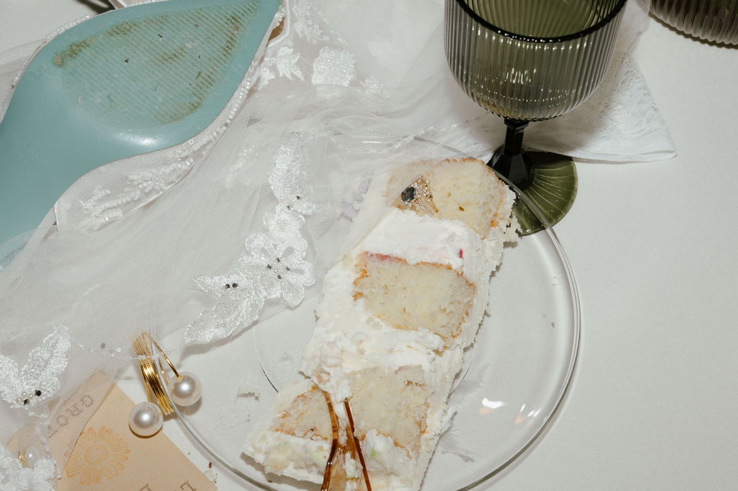 Close-up of a glass plate with wedding cake slice, lace veil, pearl earrings, and decorative glassware on white table.