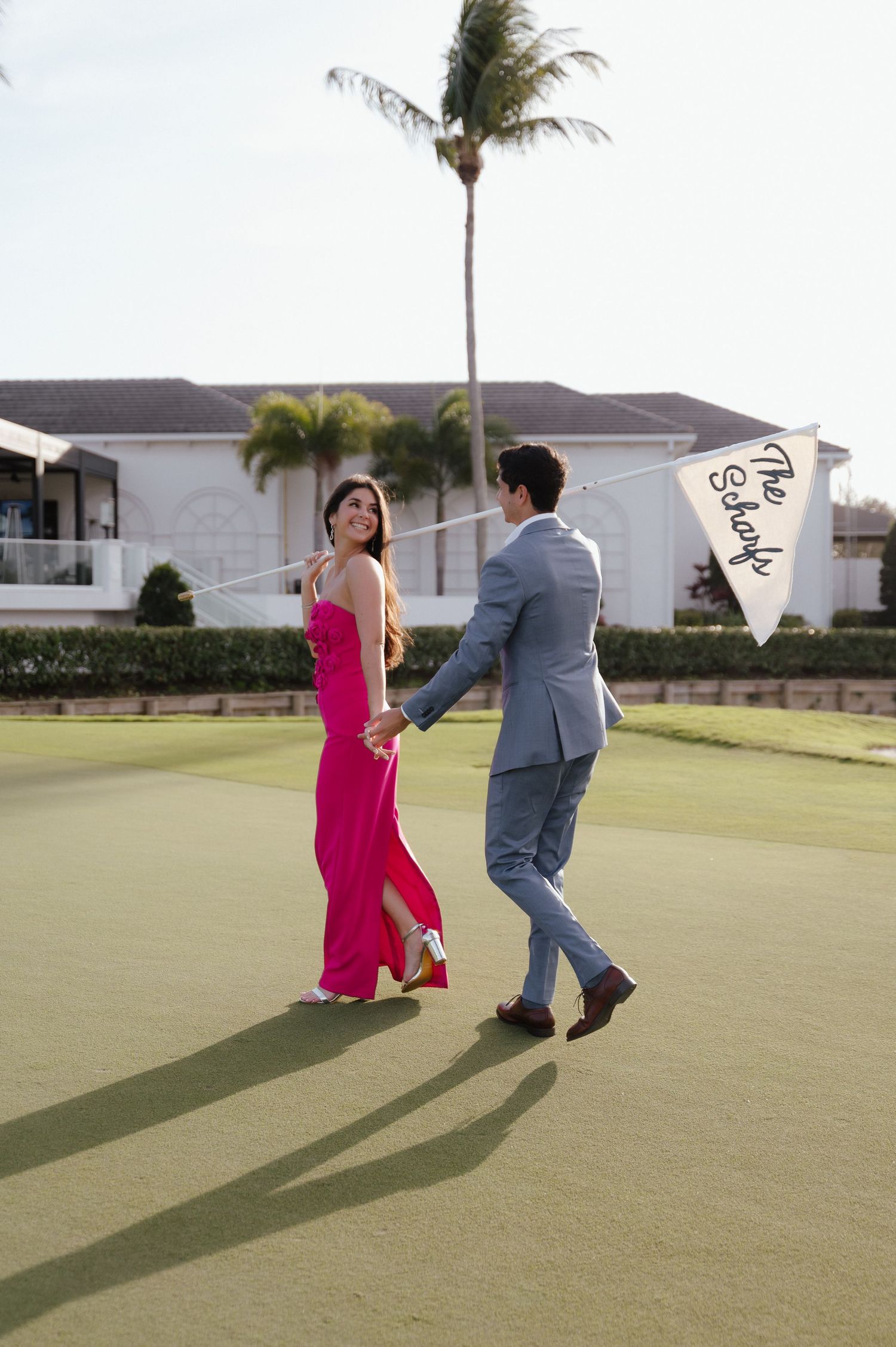 Playful couple in pink dress and grey suit running on golf course.