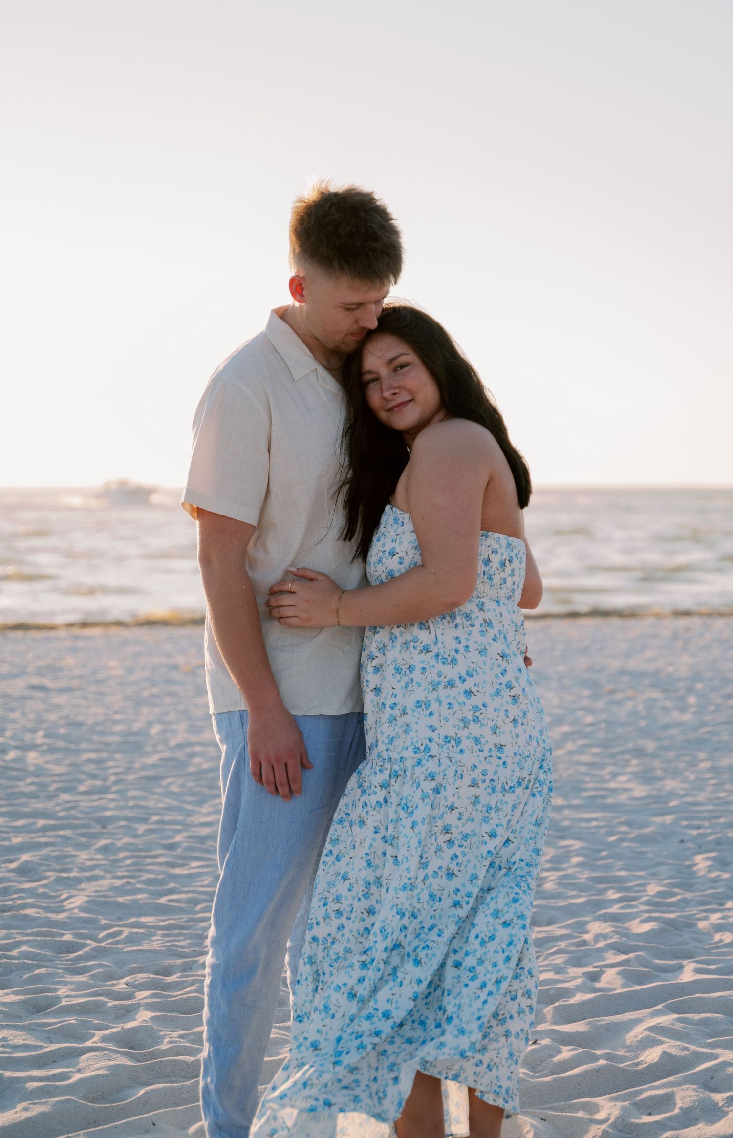 A couple embraces on a sandy beach at sunset, with the woman wearing a flowing blue floral dress.