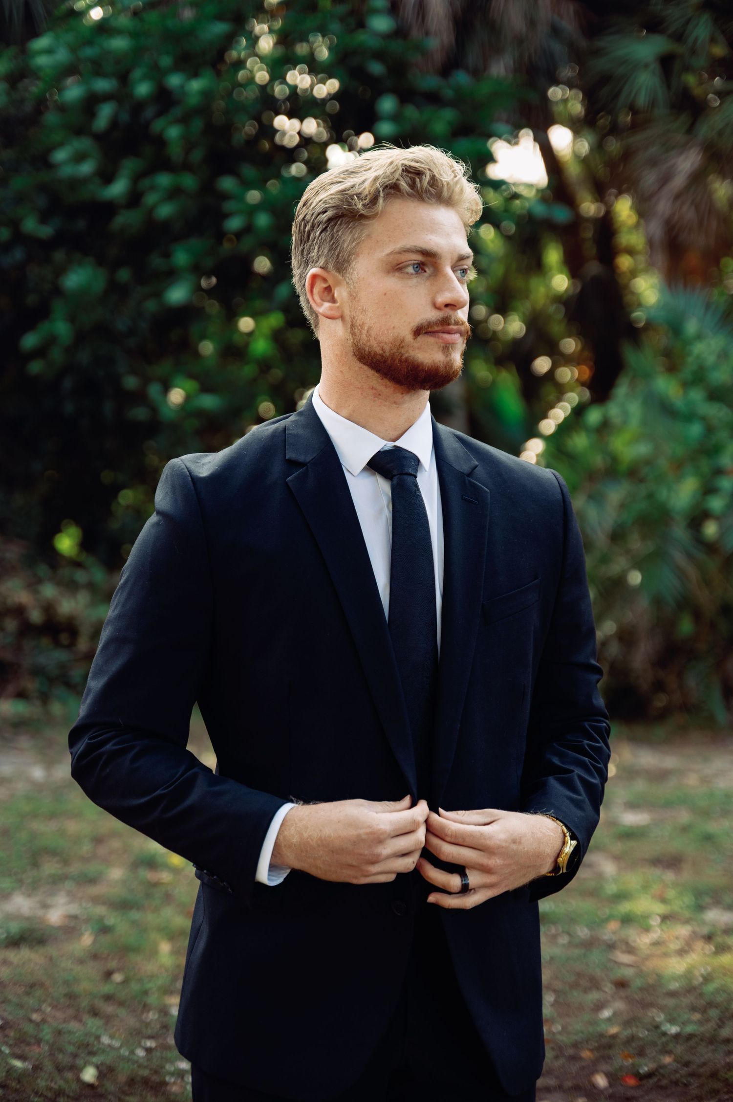 Groom adjusts suit jacket while standing in a garden setting with natural lighting.