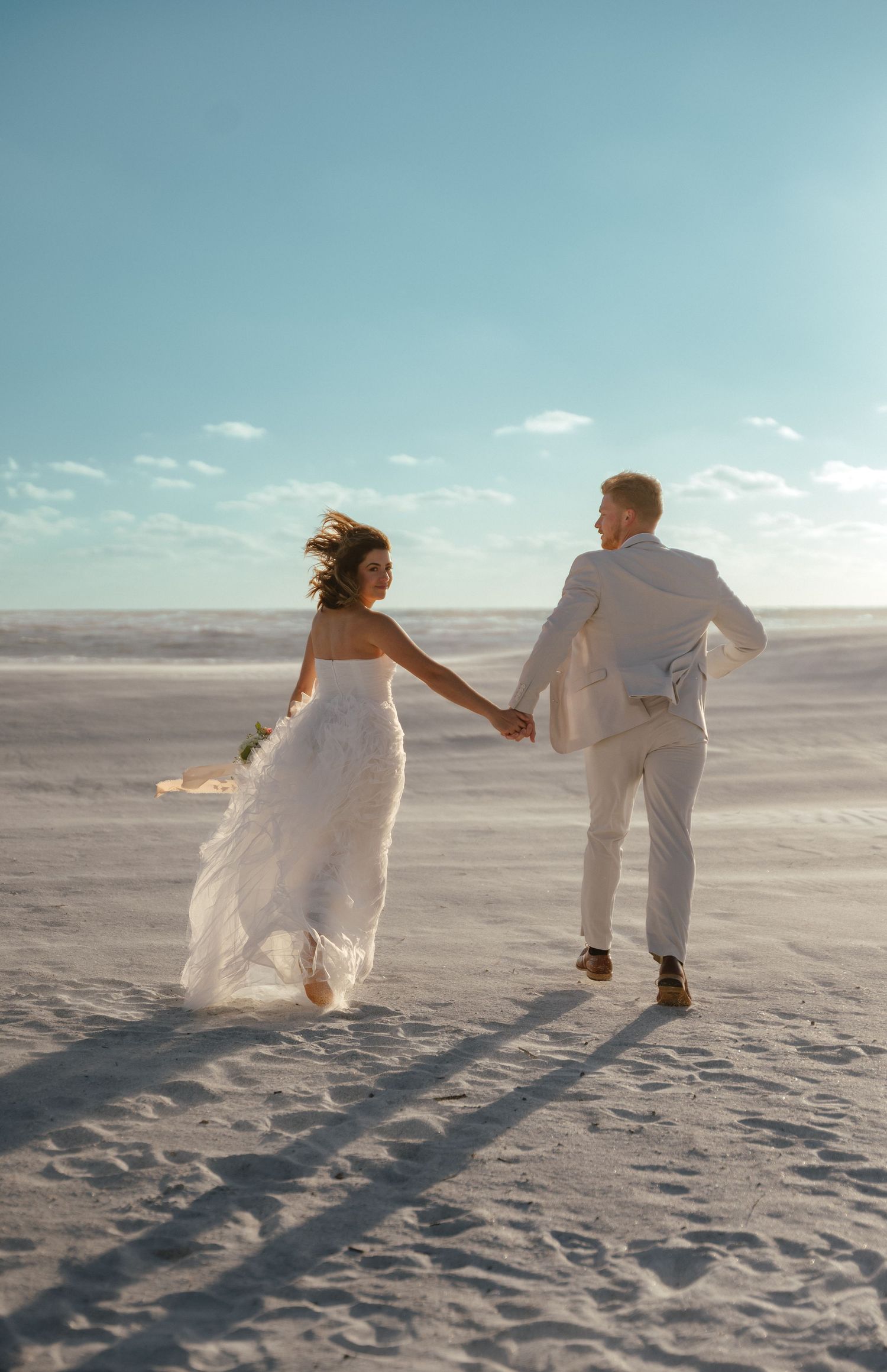 Couple in white attire running playfully along beach with long shadows.