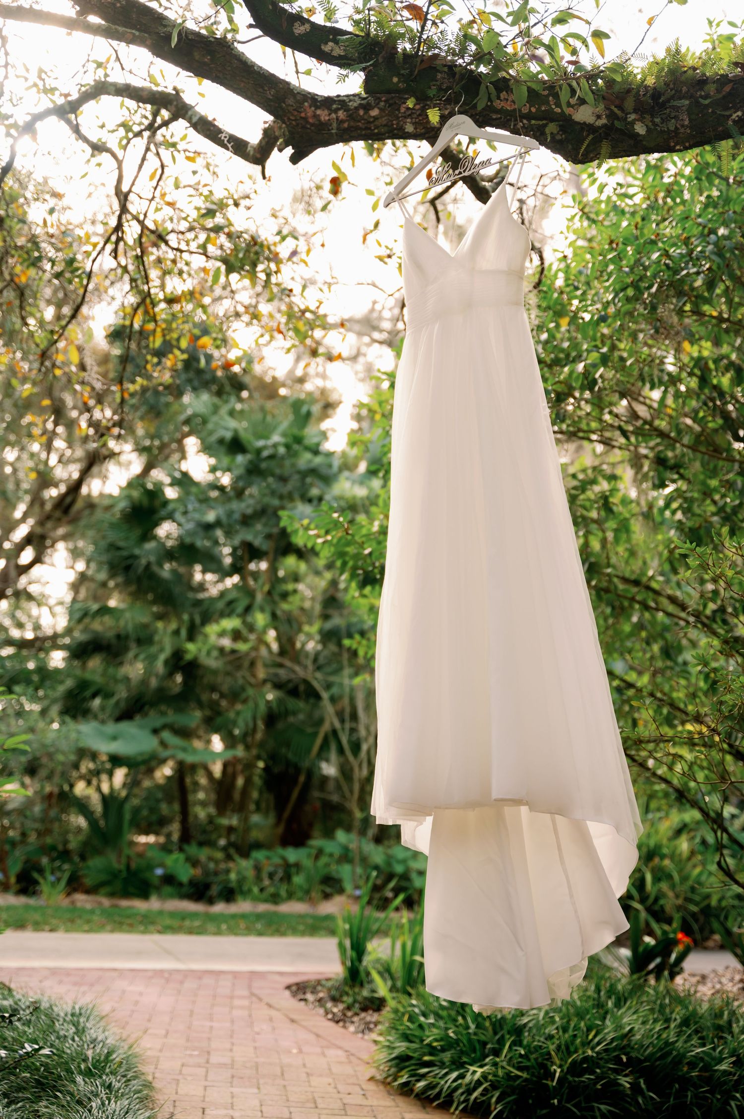 Elegant white wedding dress hanging from tree branch in a garden setting.