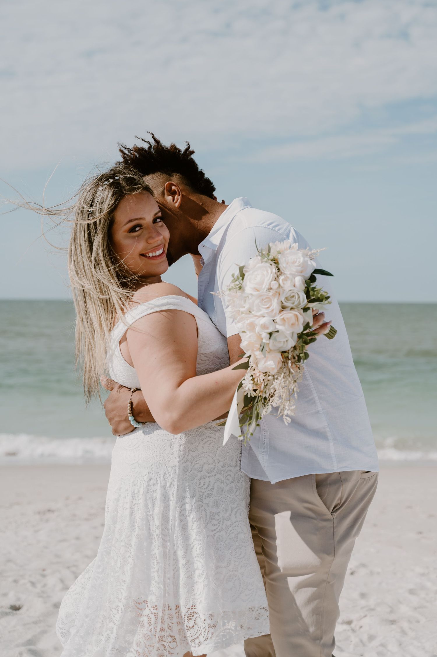 Newlyweds share intimate moments on sandy beach with white bouquet.