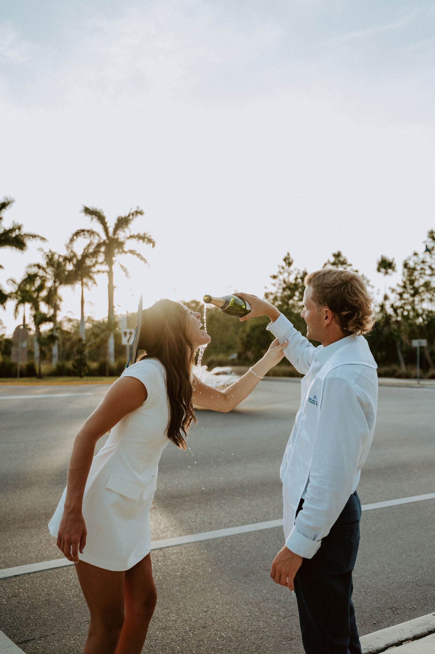 Two people playfully touch graduation caps in a palm tree-lined parking lot.