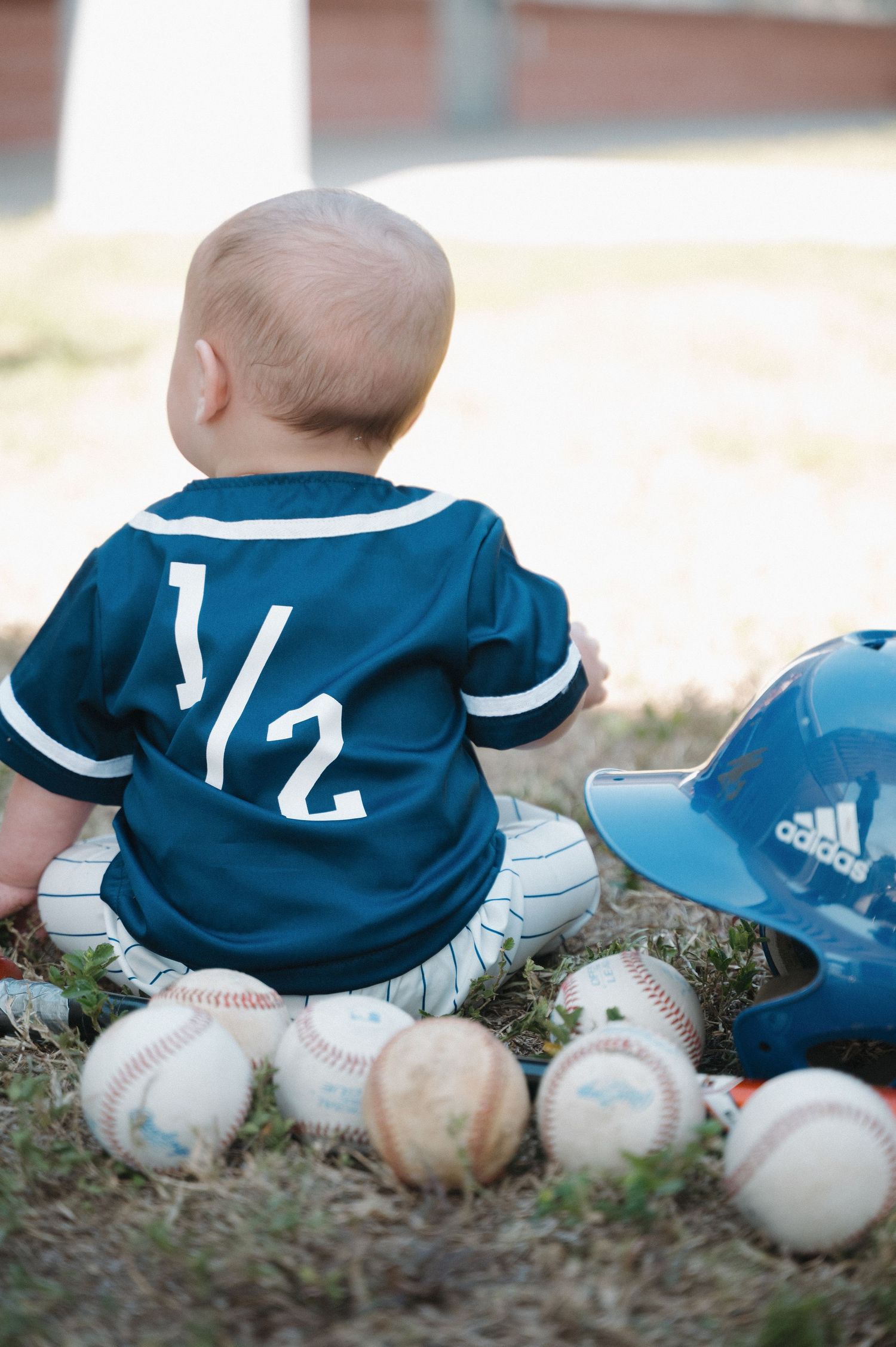 Small child wearing a baseball jersey number 1/2 sits among scattered baseballs and baseball gear.