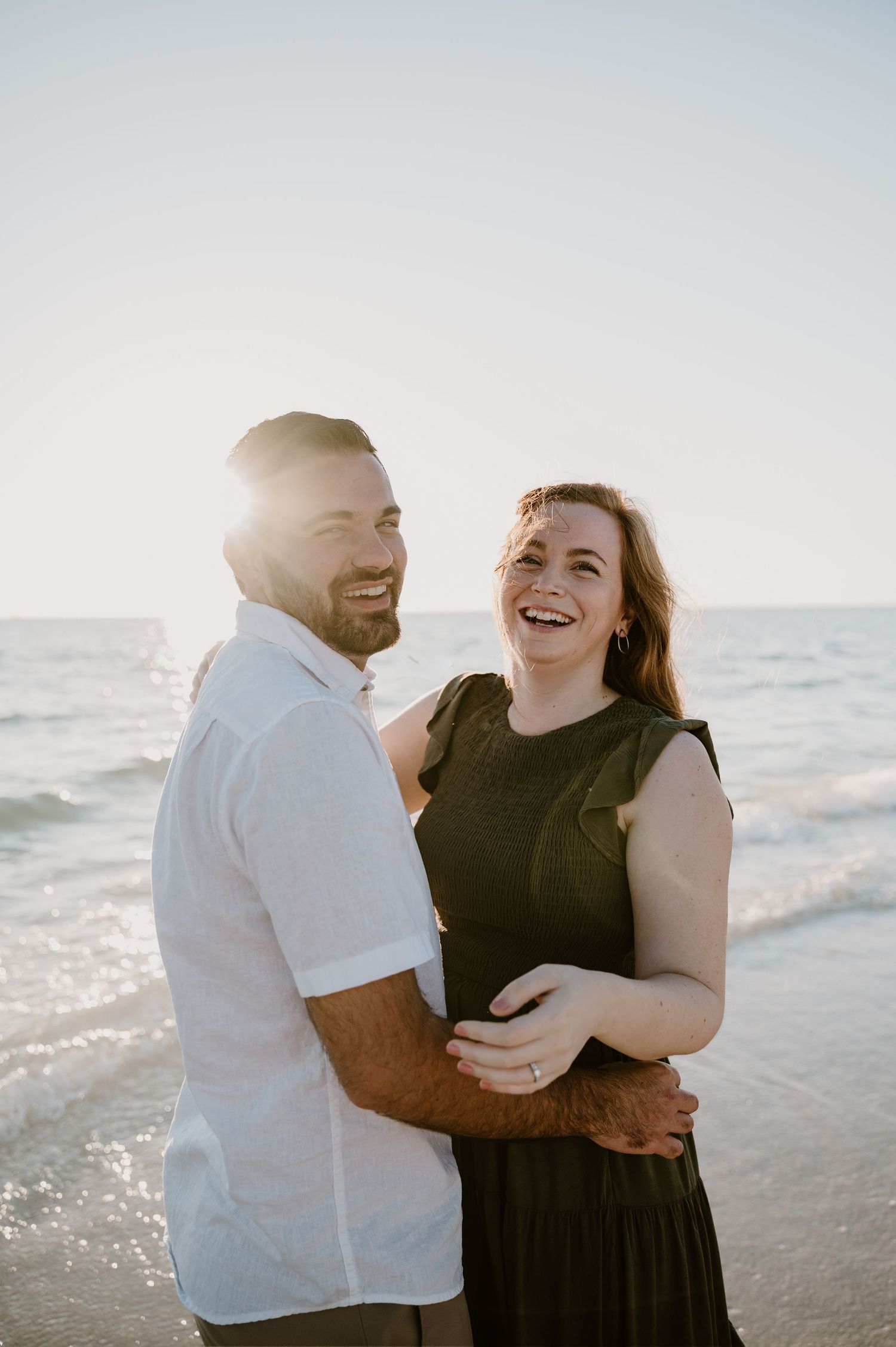 Happy couple laughing together on beach during golden hour with ocean waves behind them.
