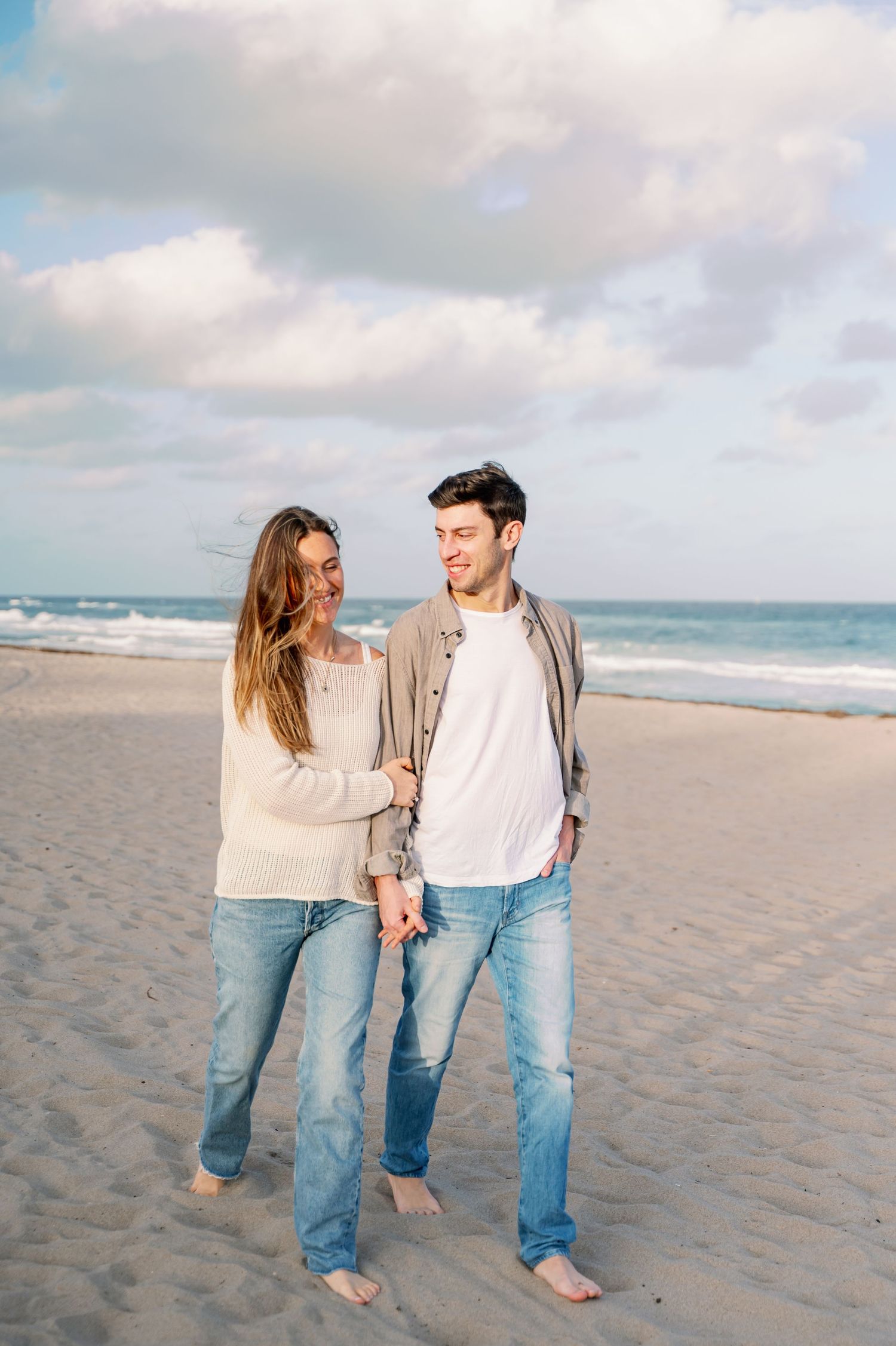A couple in casual clothing stands barefoot on a sandy beach with ocean waves and cloudy sky in the background.