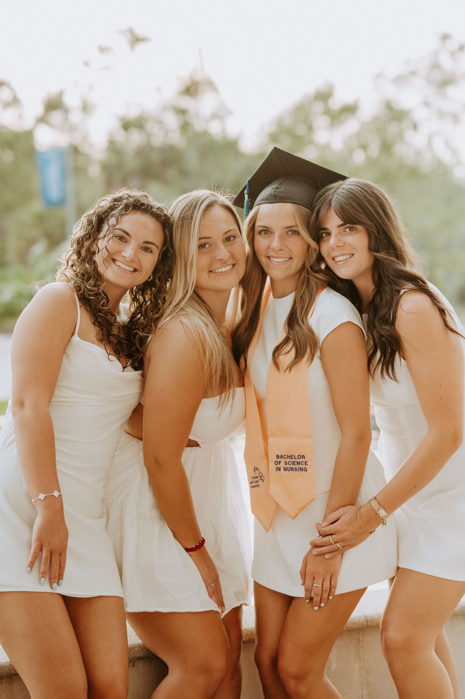 Four graduates in white dresses celebrate together on a sunny day.