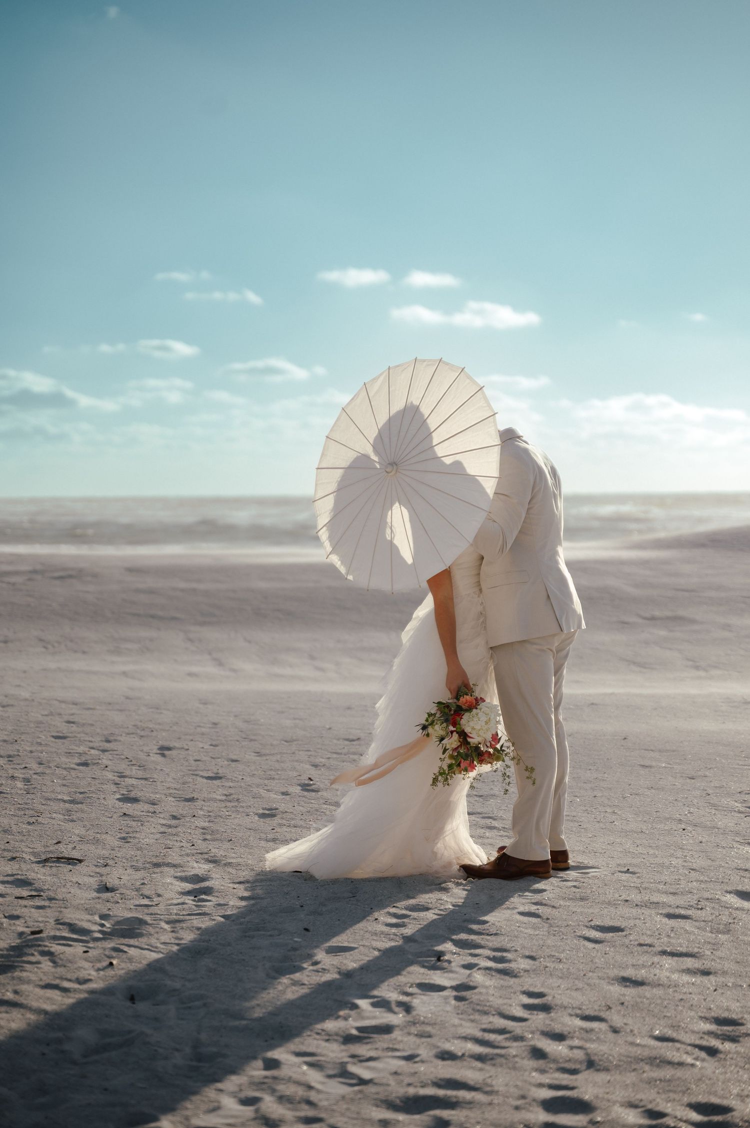 Romantic beach wedding photos with white parasols and flowing dress on sandy shore.