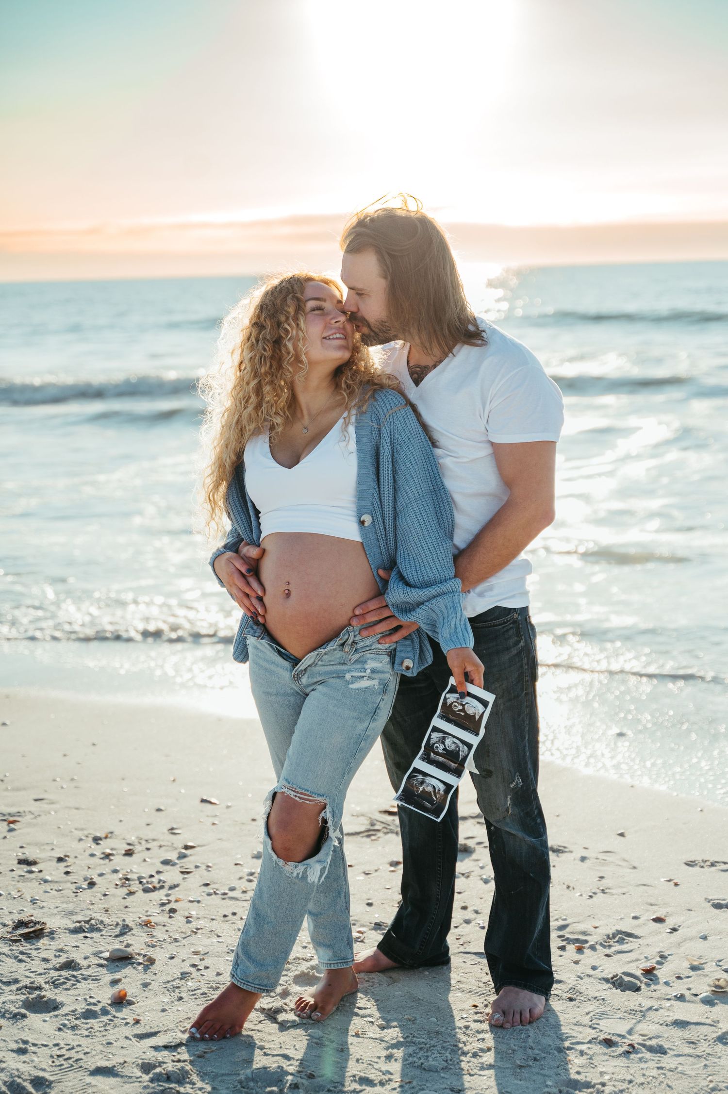 Expecting couple sharing intimate moment on beach with ultrasound photos.