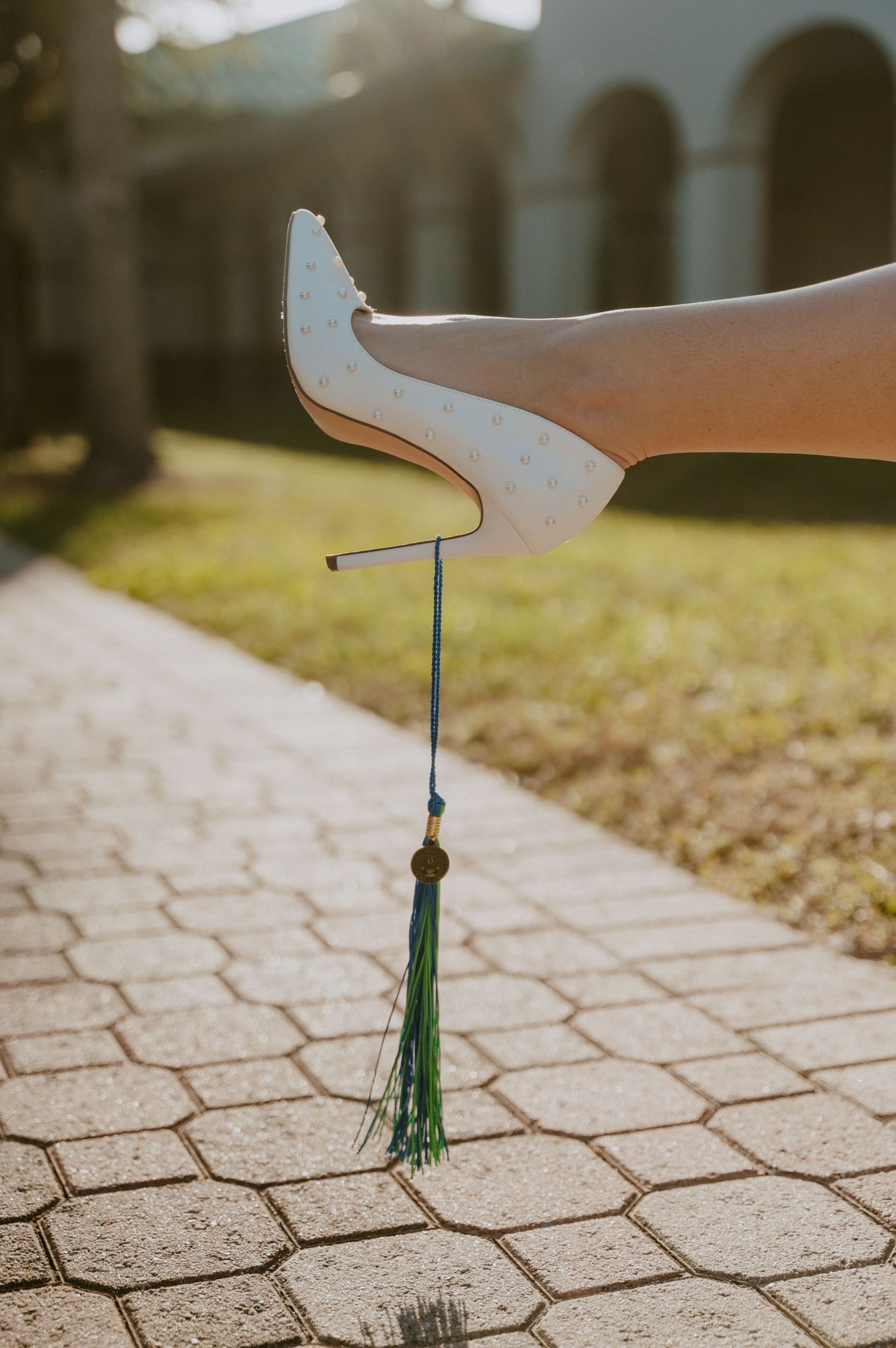 White high heel shoe with green tassel against stone pavement in sunlight.