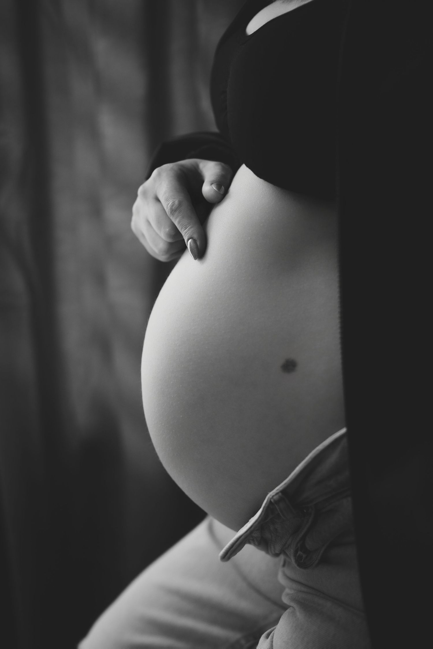 Black and white photo of a pregnant woman cradling her bare baby bump, wearing a dark bra and white pants.