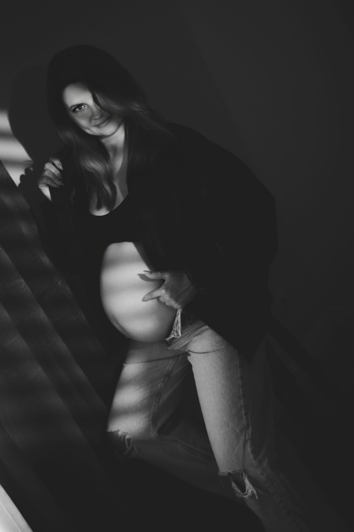 Black and white photo of a pregnant woman in black top and denim shorts posing on stairs with dramatic lighting.