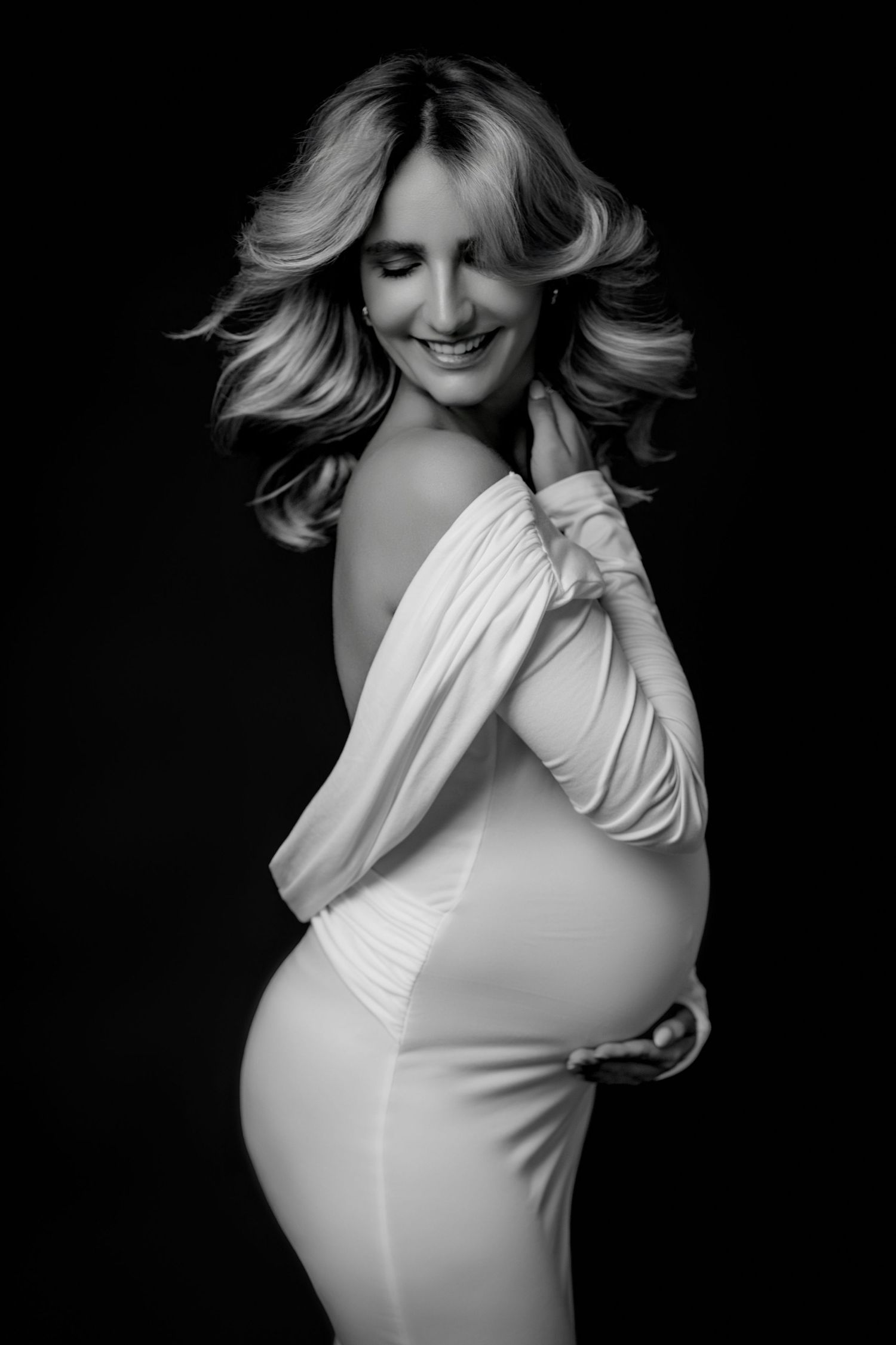 Black and white portrait of a smiling pregnant woman in a white off-shoulder gown, showcasing her baby bump.
