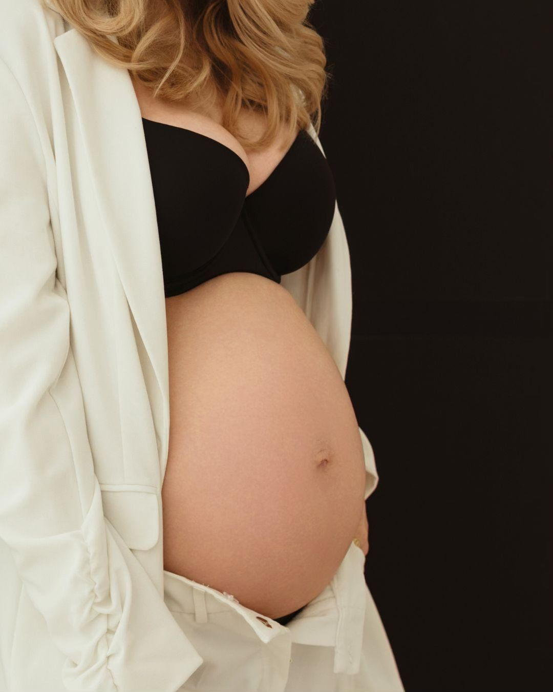 A side view of a pregnant belly with a white open cardigan against a black background.