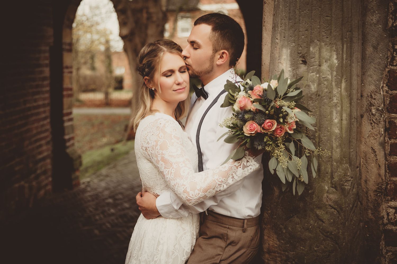 Romantic wedding couple poses against rustic brick wall with floral bouquet.