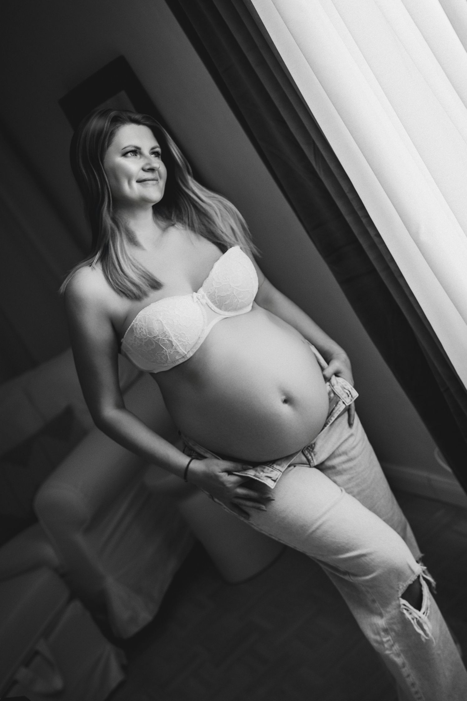 Black and white photo of a smiling pregnant woman in a white bra and jeans standing by a window.