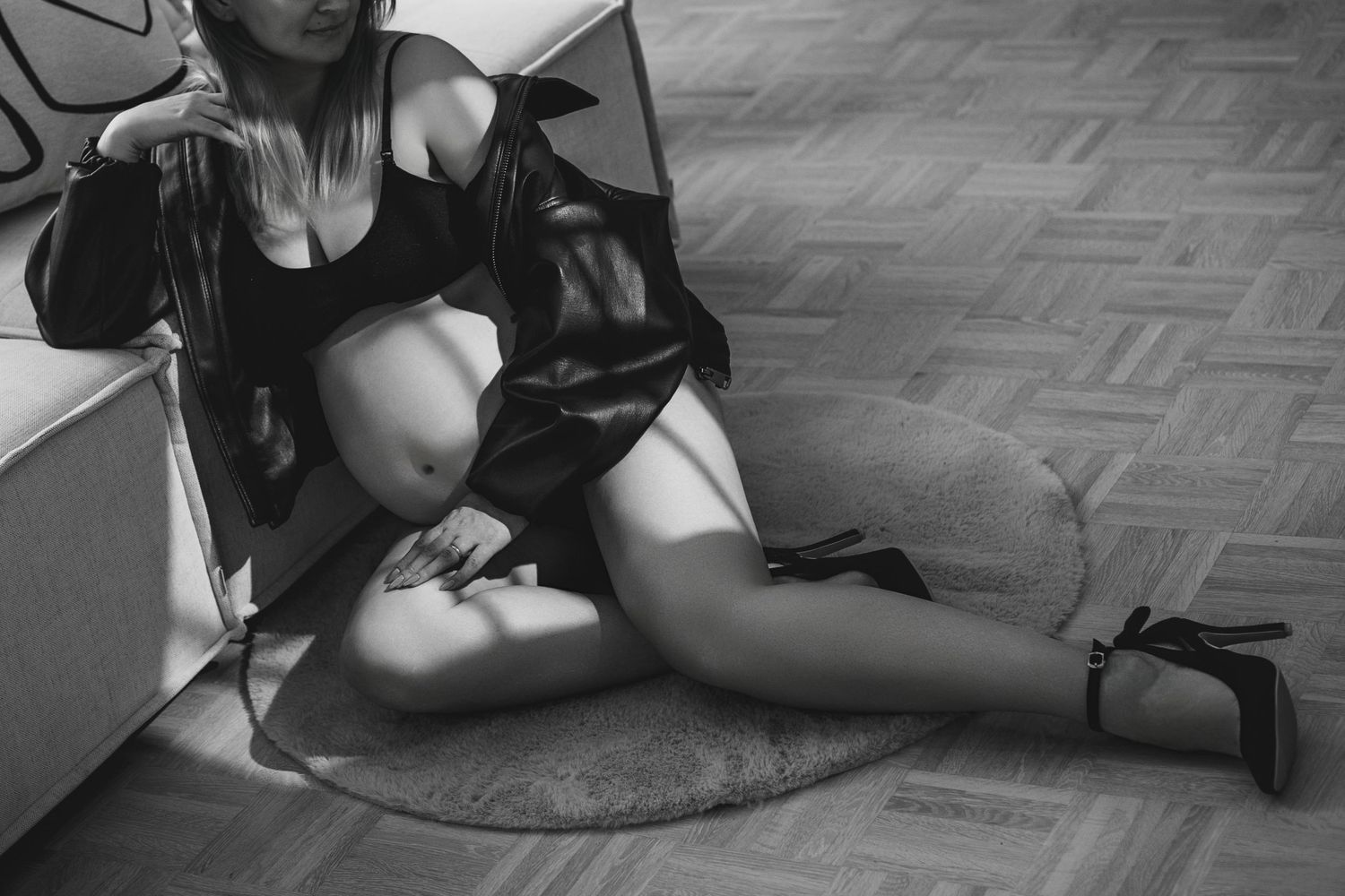 Black and white artistic photo of a woman in an open jacket sitting on a floor with crossed legs wearing heels.