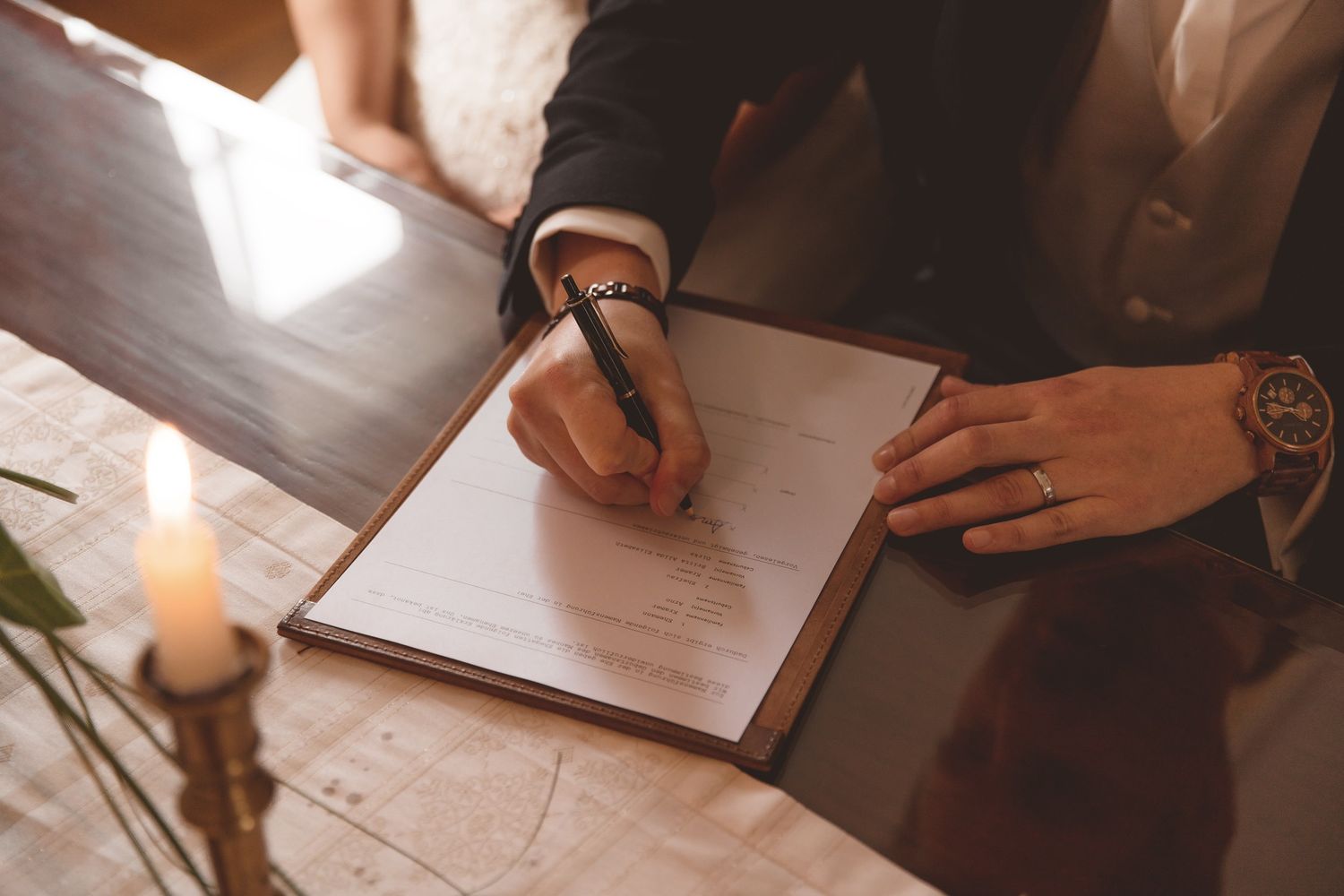 Person signing legal documents with pen at wooden desk while another person watches and provides guidance.
