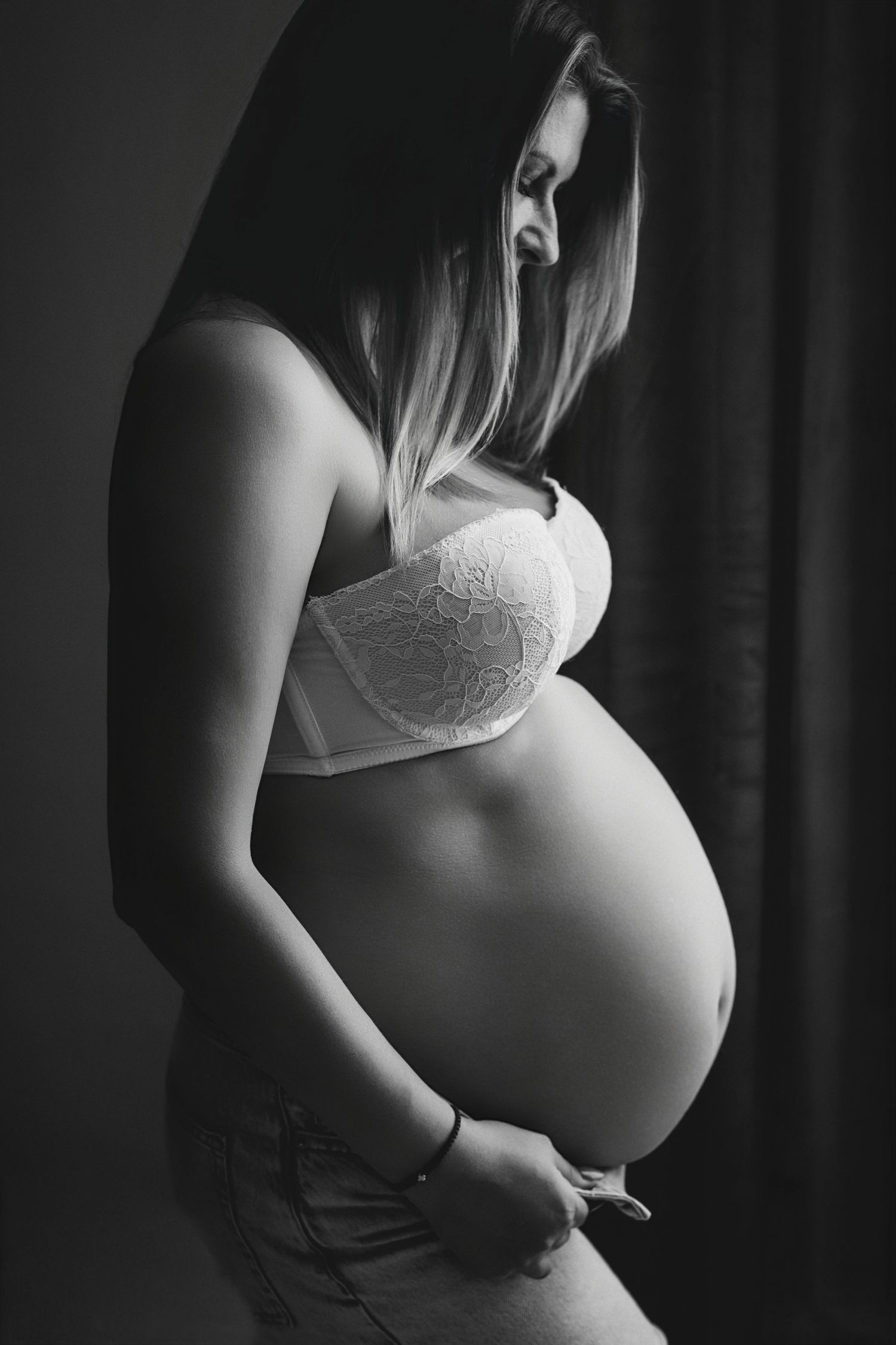 Black and white photo of a pregnant woman in lace bra cradling her baby bump in a studio setting.