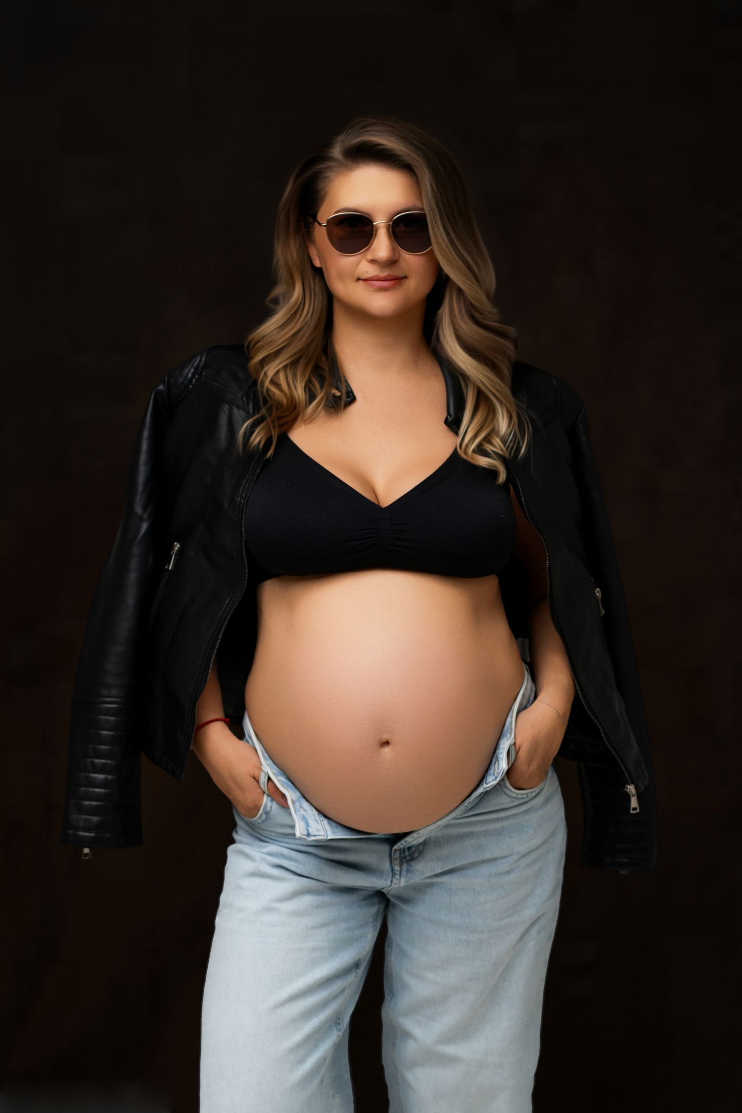 Pregnant woman in black crop top, leather jacket, jeans, and sunglasses posing confidently against dark background.