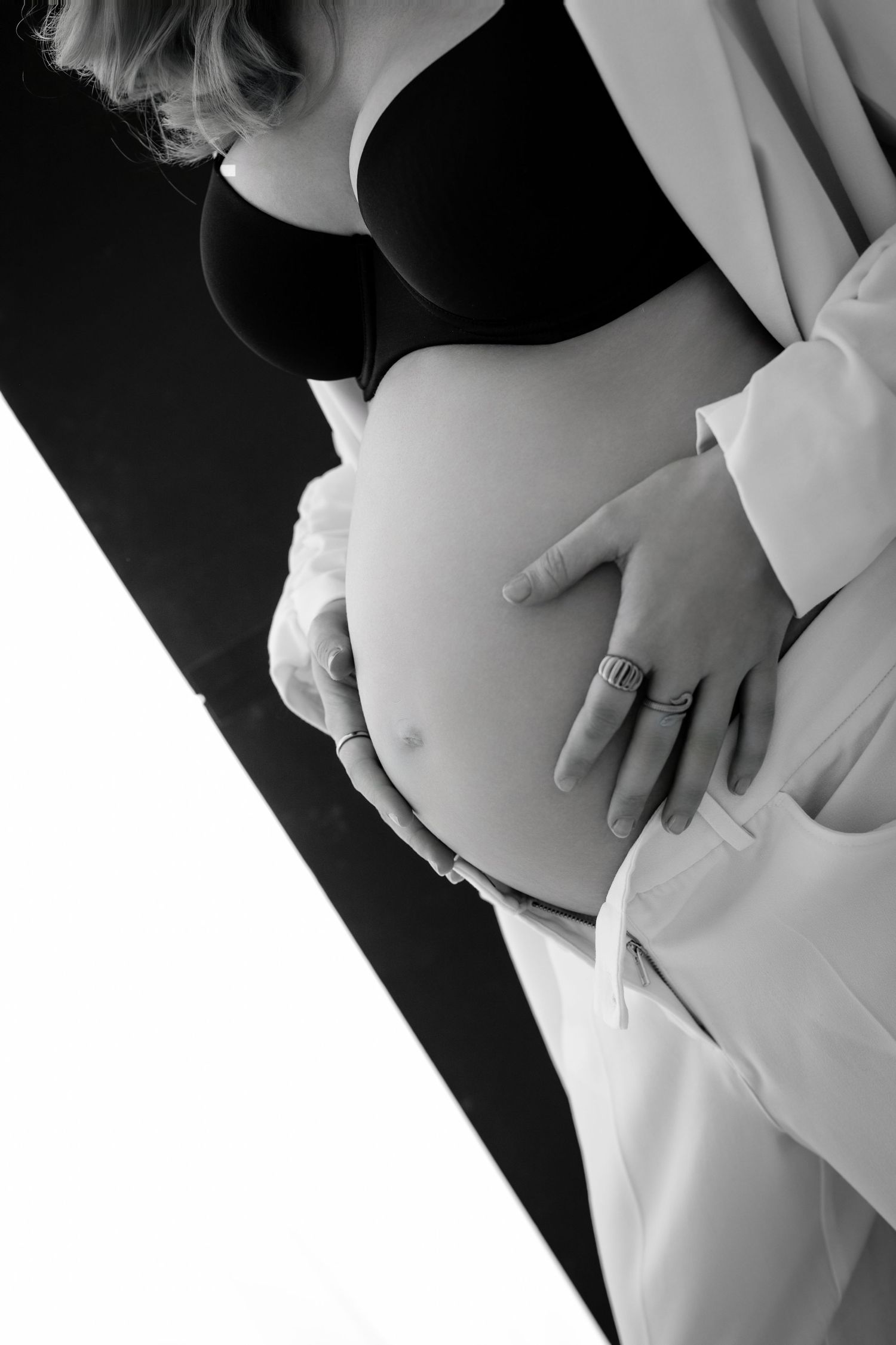 Artistic black and white maternity photo showing pregnant belly with hands wearing ring against light and dark contrast.