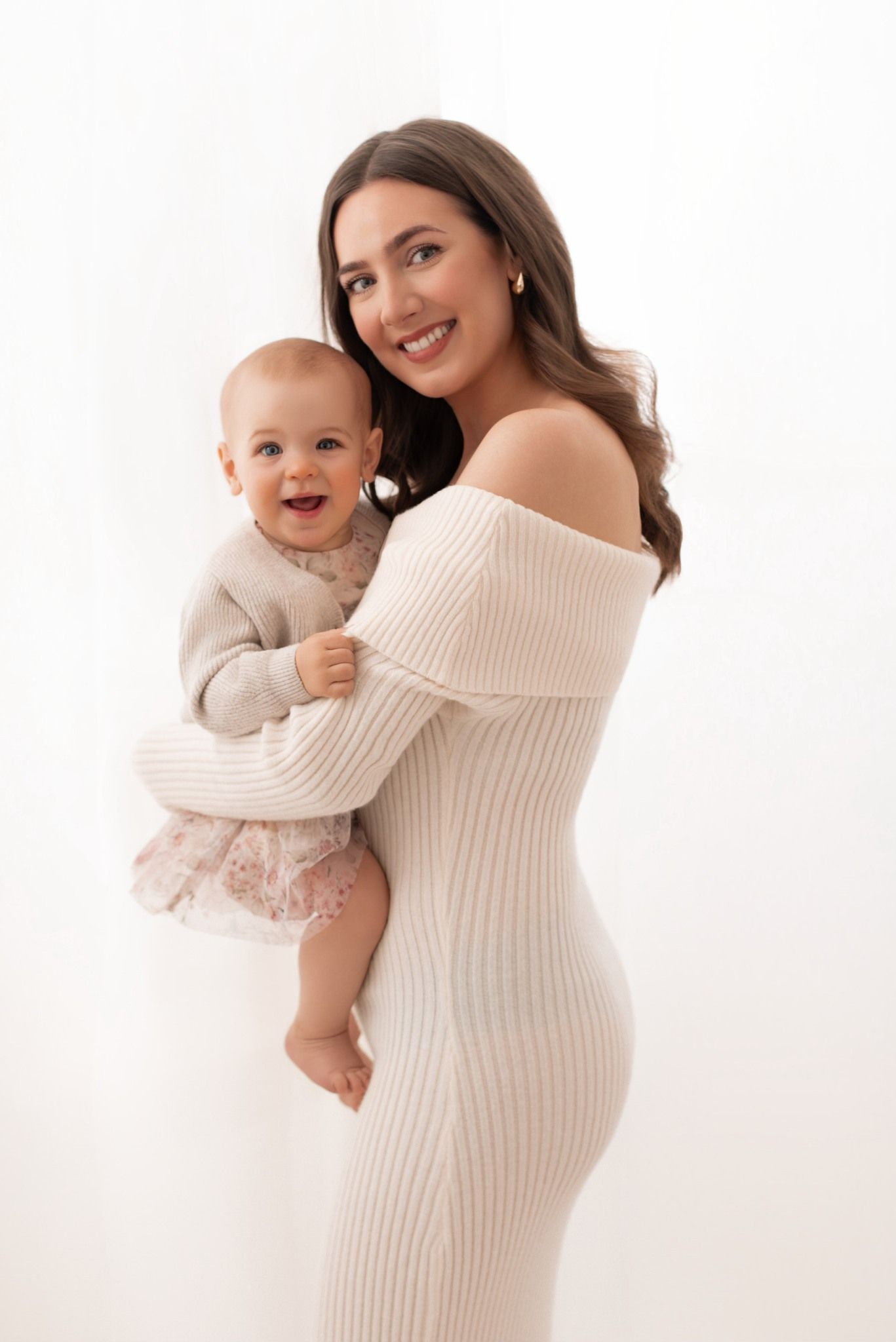 A mother in a cream sweater holds a baby wearing matching cream clothing against a white studio backdrop.