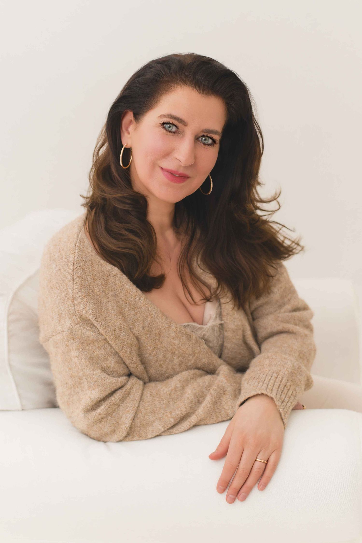 A person with dark hair wearing a cozy beige sweater poses while reclining on white bedding.