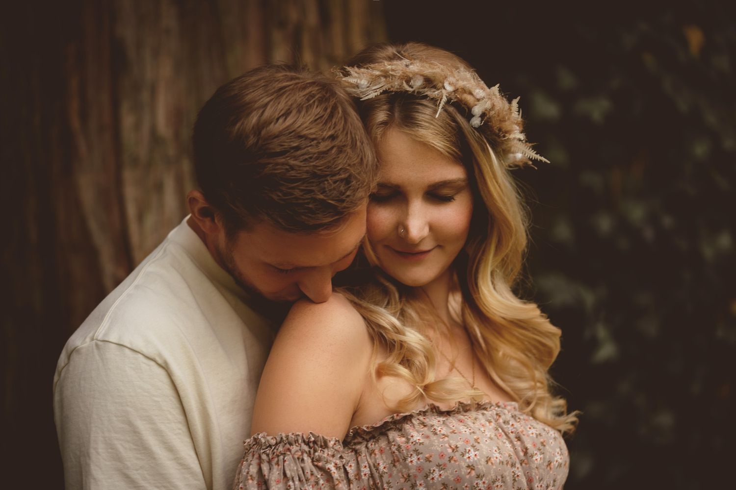 Romantic couple embraces tenderly outdoors, with bride wearing floral crown and off-shoulder dress in golden sunset light.