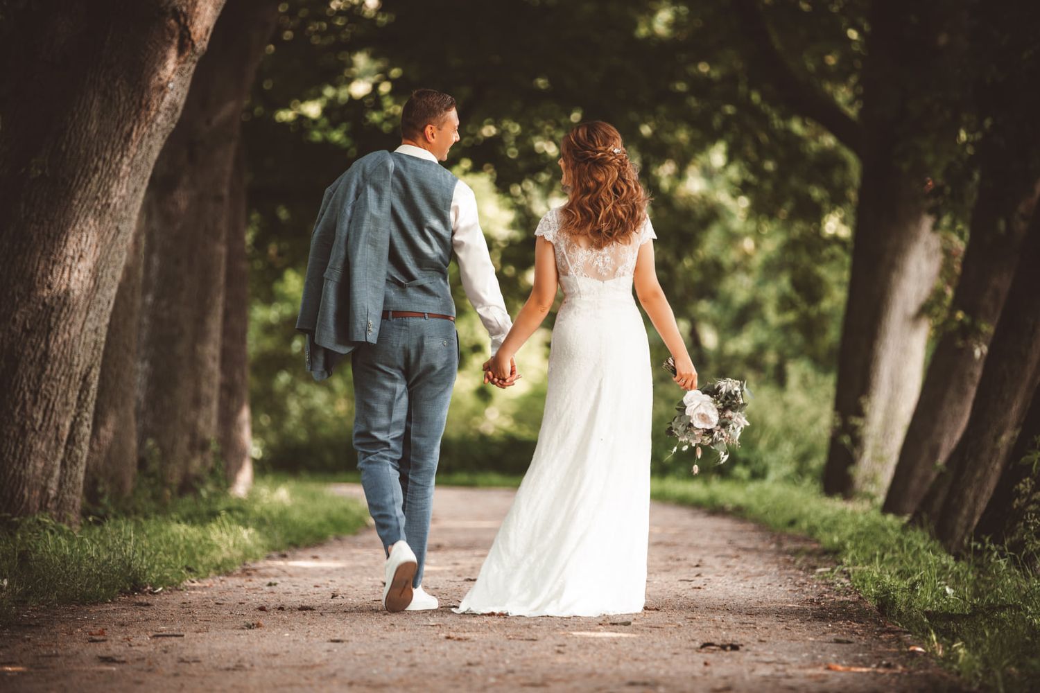 Wedding couple walks hand in hand down tree-lined path in romantic garden setting.