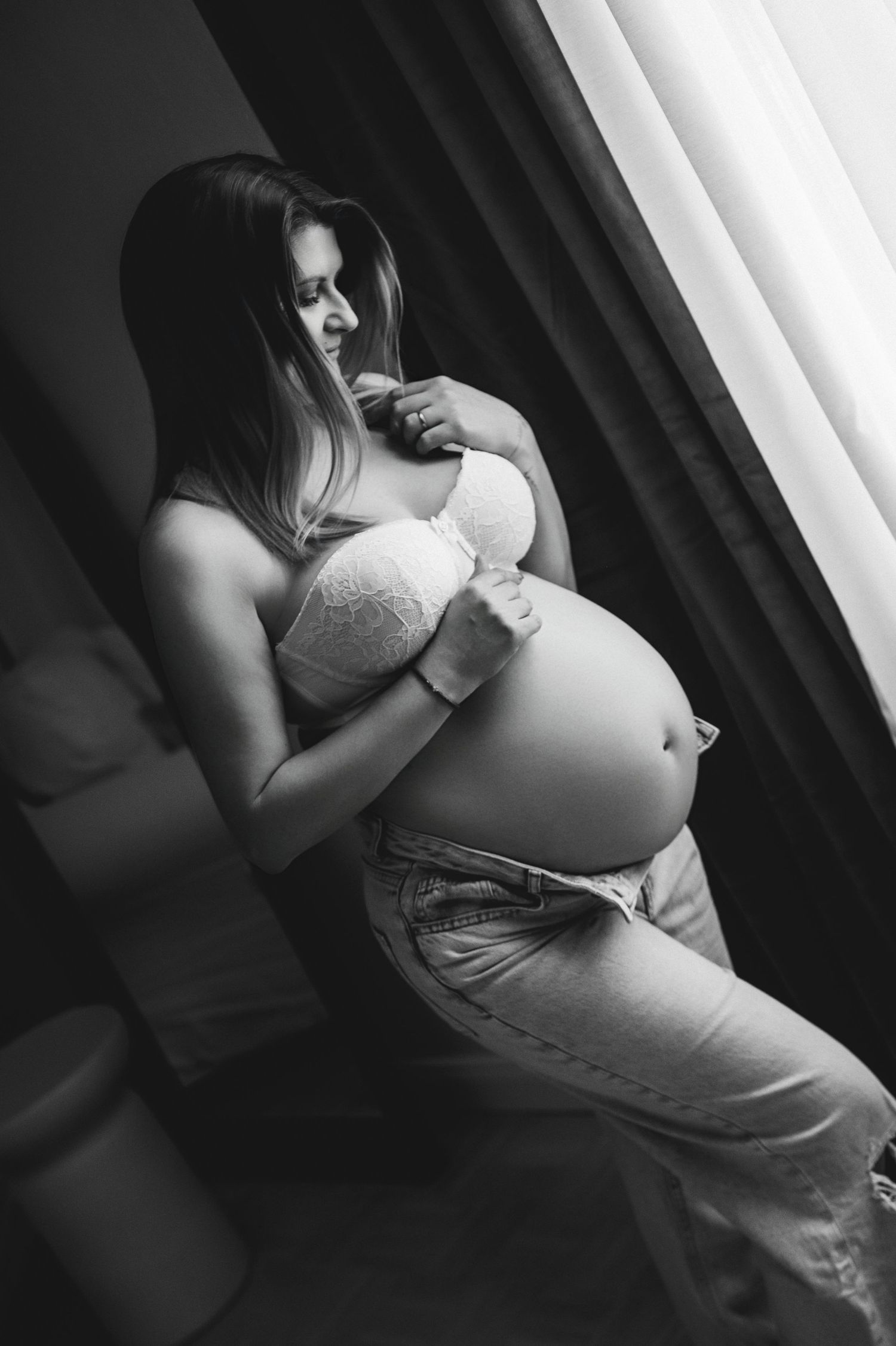 Black and white photo of a pregnant woman in a bra and jeans cradling her baby bump by a window.