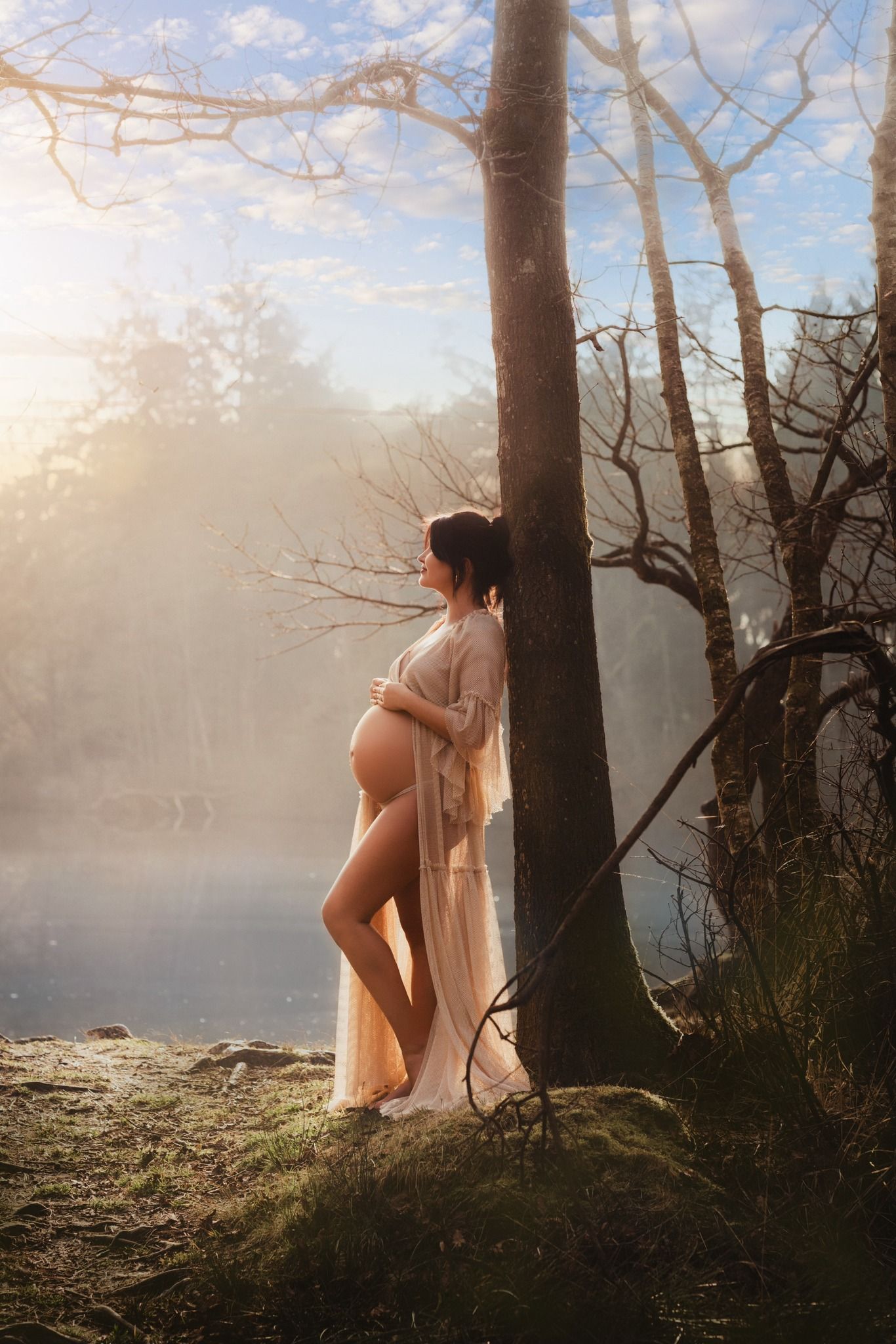 Pregnant woman in sheer robe leaning against tree by misty lake at golden hour sunrise in nature.