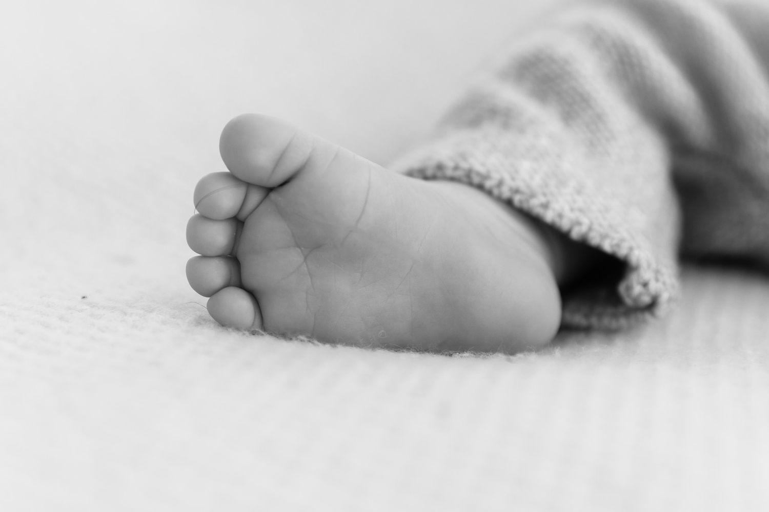 A close-up black and white photograph of tiny baby feet with delicate toes against a soft textured background.