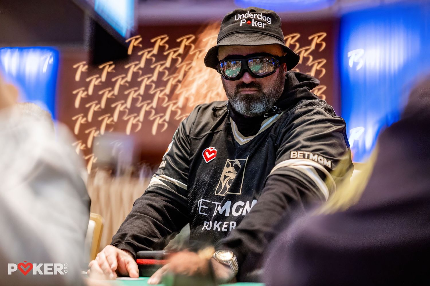 A poker player wearing sunglasses and a black jacket sits at a poker table during a professional tournament.