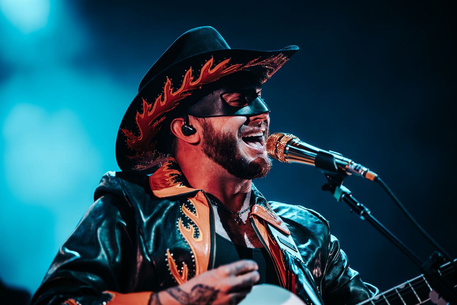 Concert performer in leather jacket and cowboy hat singing passionately into microphone with blue stage lighting.