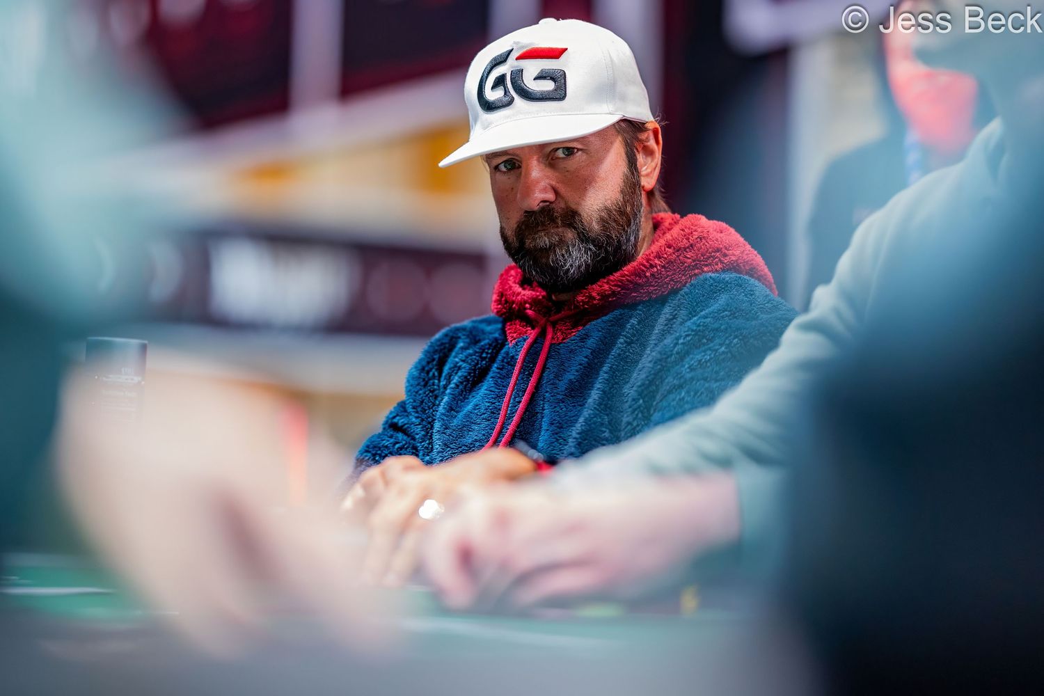 Bearded man wearing a GG poker white cap sits focused at a poker table during a professional tournament.