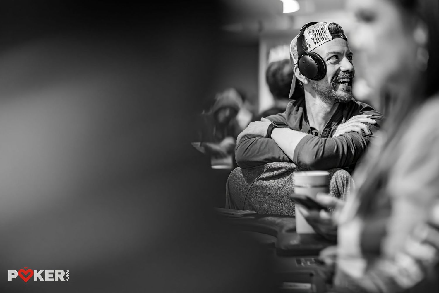 A smiling man wearing headphones relaxes at a poker table during a tournament, captured in black and white photography.