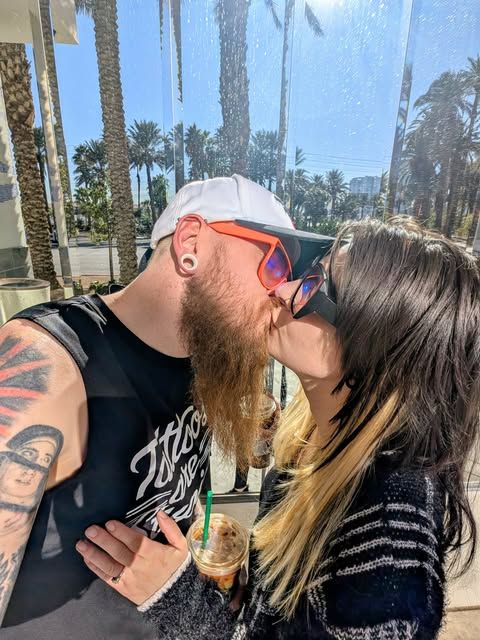 A couple shares a kiss while holding drinks outdoors with palm trees in the background on a sunny day.