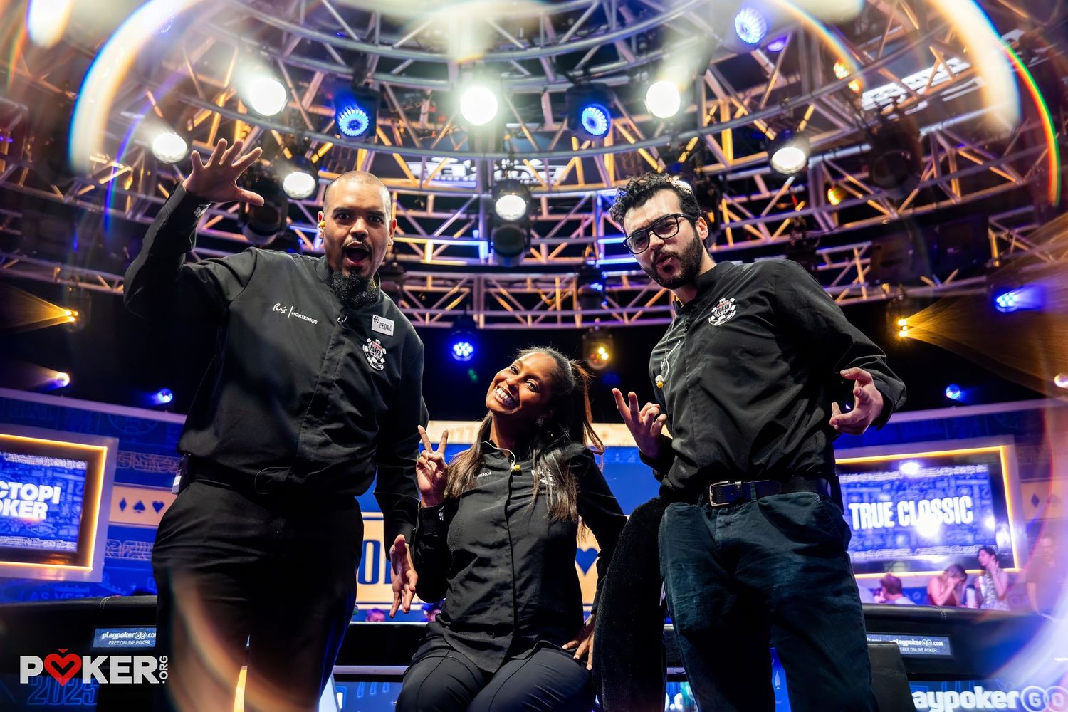 Three people celebrate joyfully on a poker tournament stage with bright lights and signage in the background.