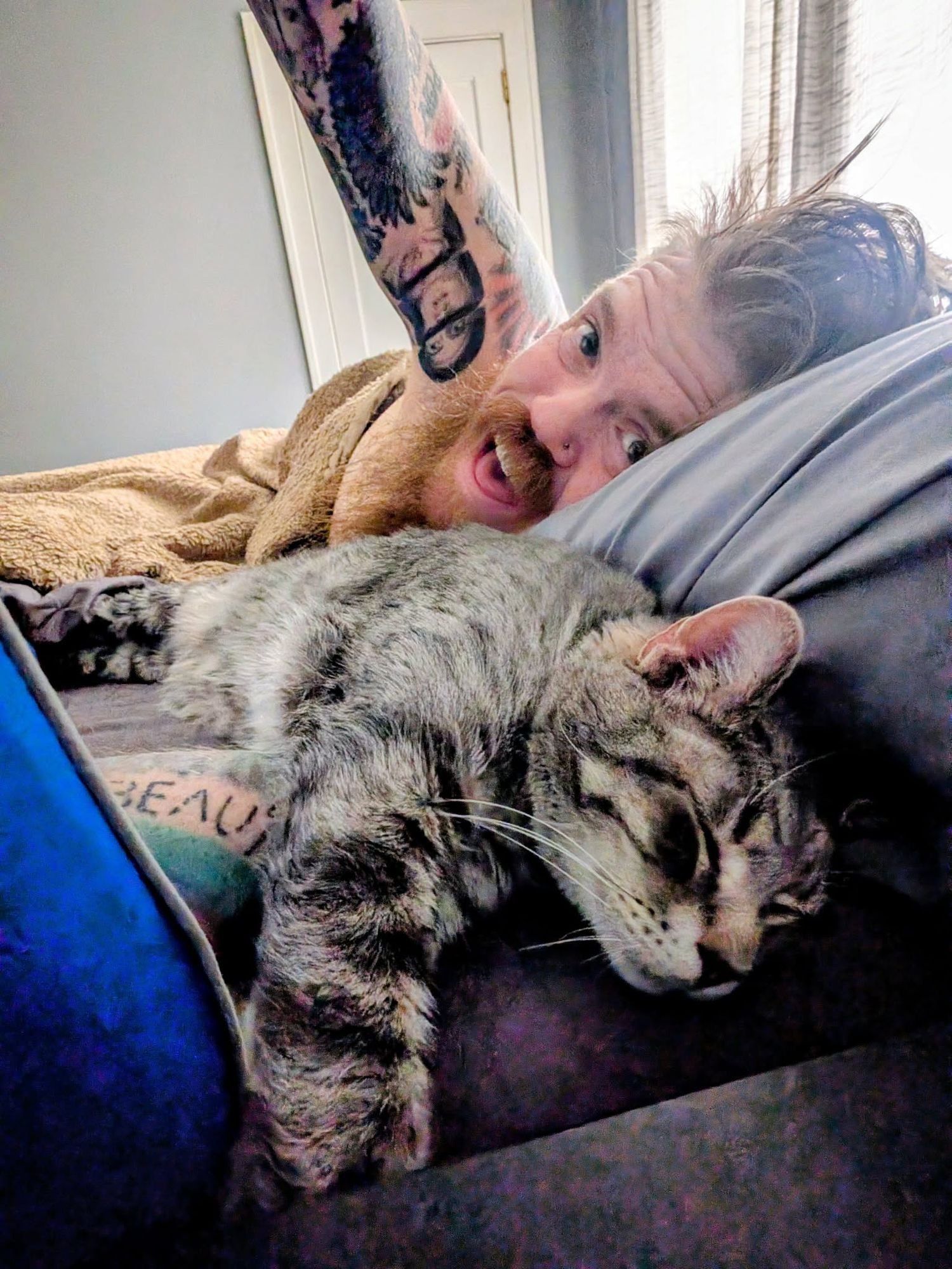 A gray tabby cat sleeps contentedly on a couch next to someone under a tan blanket.