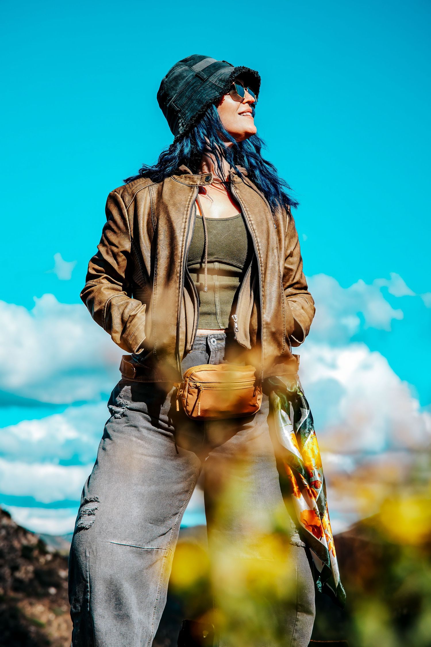 Woman with blue hair wearing a bucket hat, sunglasses, brown jacket, and fanny pack poses outdoors under a bright blue sky.