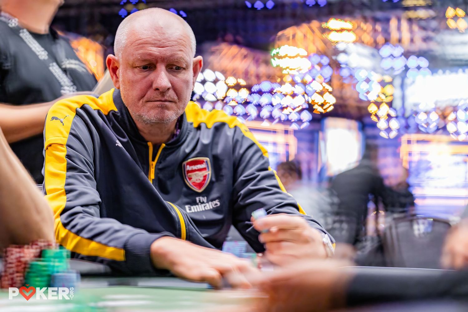 A poker player wearing a yellow and black Arsenal jacket sits at a casino table with colorful lights in the background.