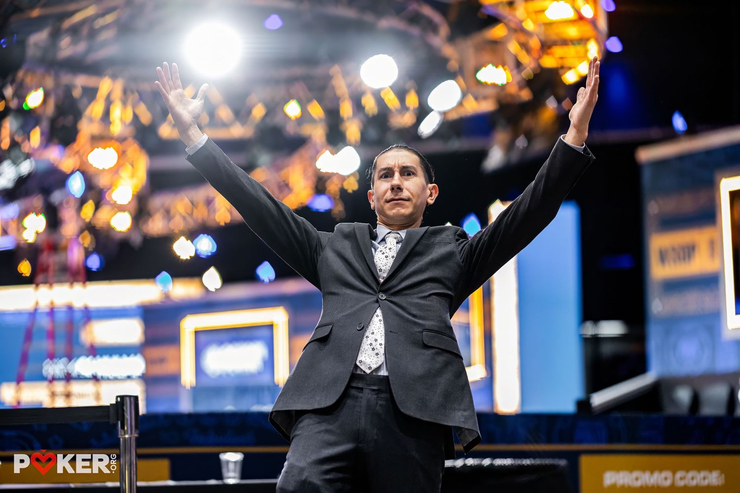 Jubilant poker player celebrates tournament victory with arms raised at gaming table under bright stage lights.