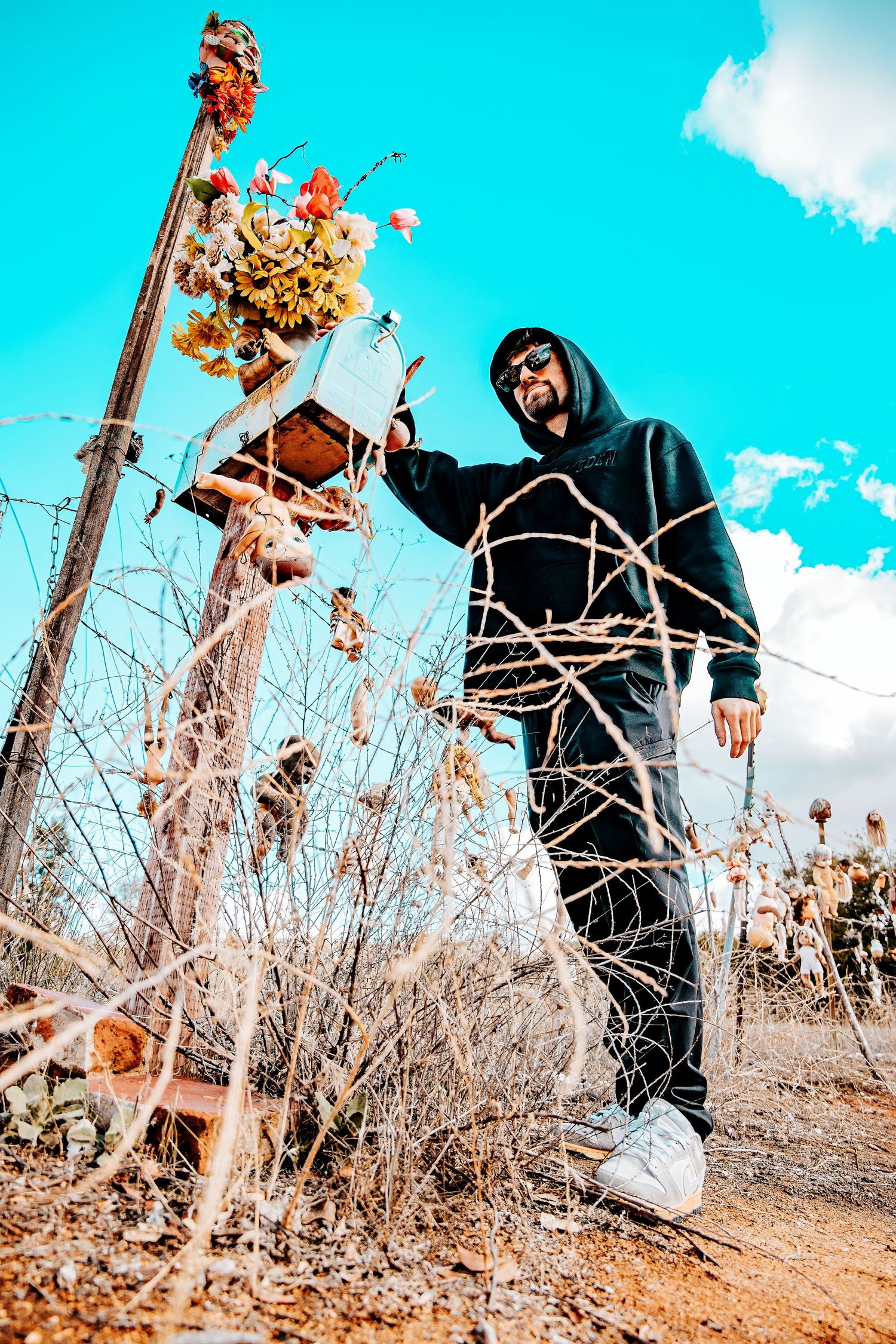 Man in black hoodie and sunglasses standing near a rustic roadside memorial with flowers under a blue sky.