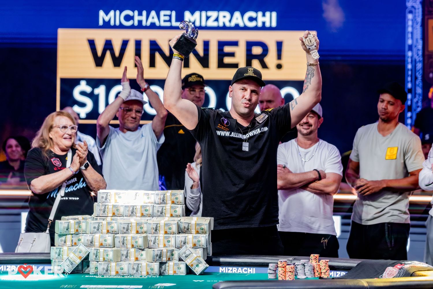 Michael Mizrachi celebrates his poker tournament win, raising arms over stacks of cash with Winner sign behind him.